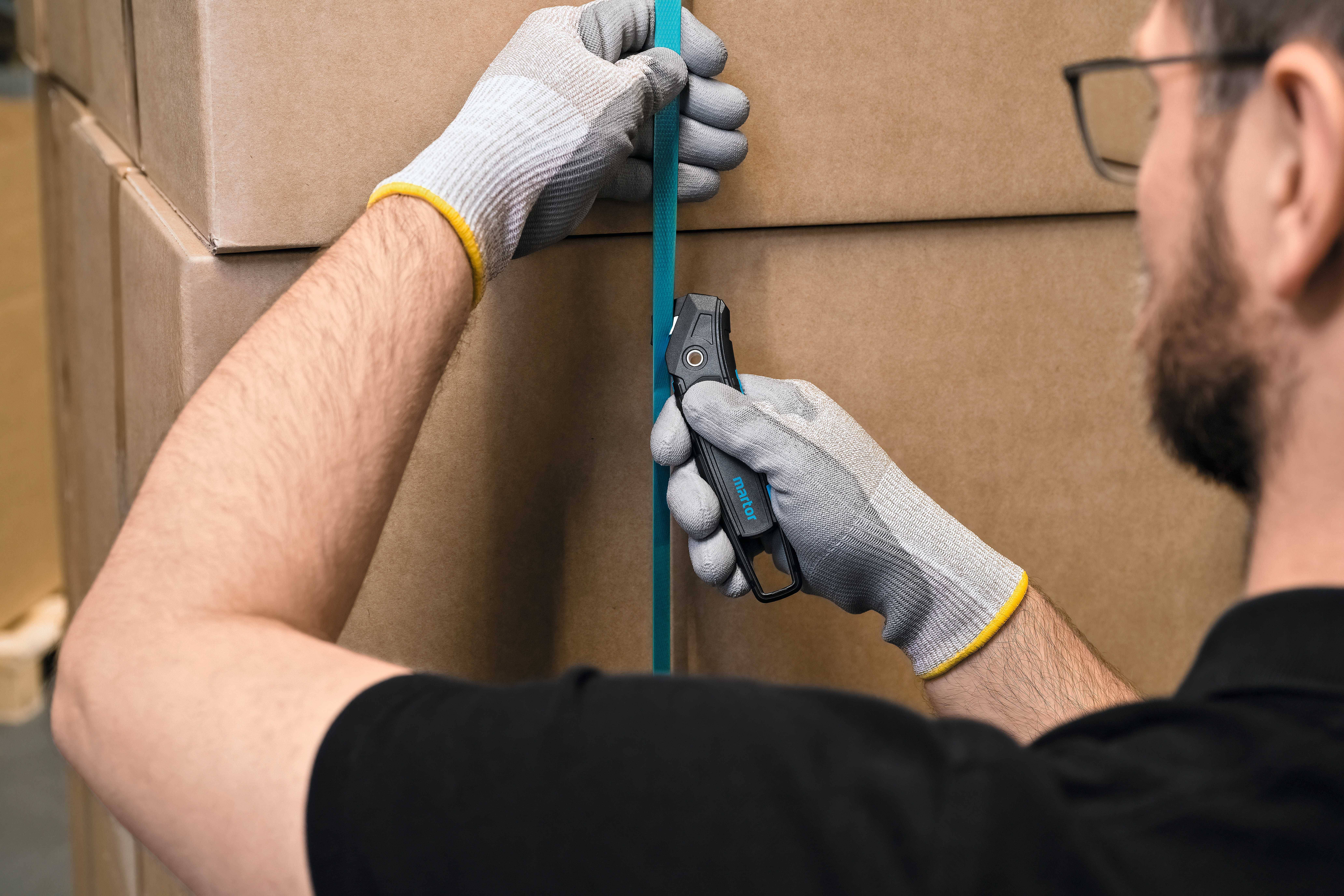 Een persoon met handschoenen snijdt met een blauw mes een kunststofband van een grote kartonnen doos.