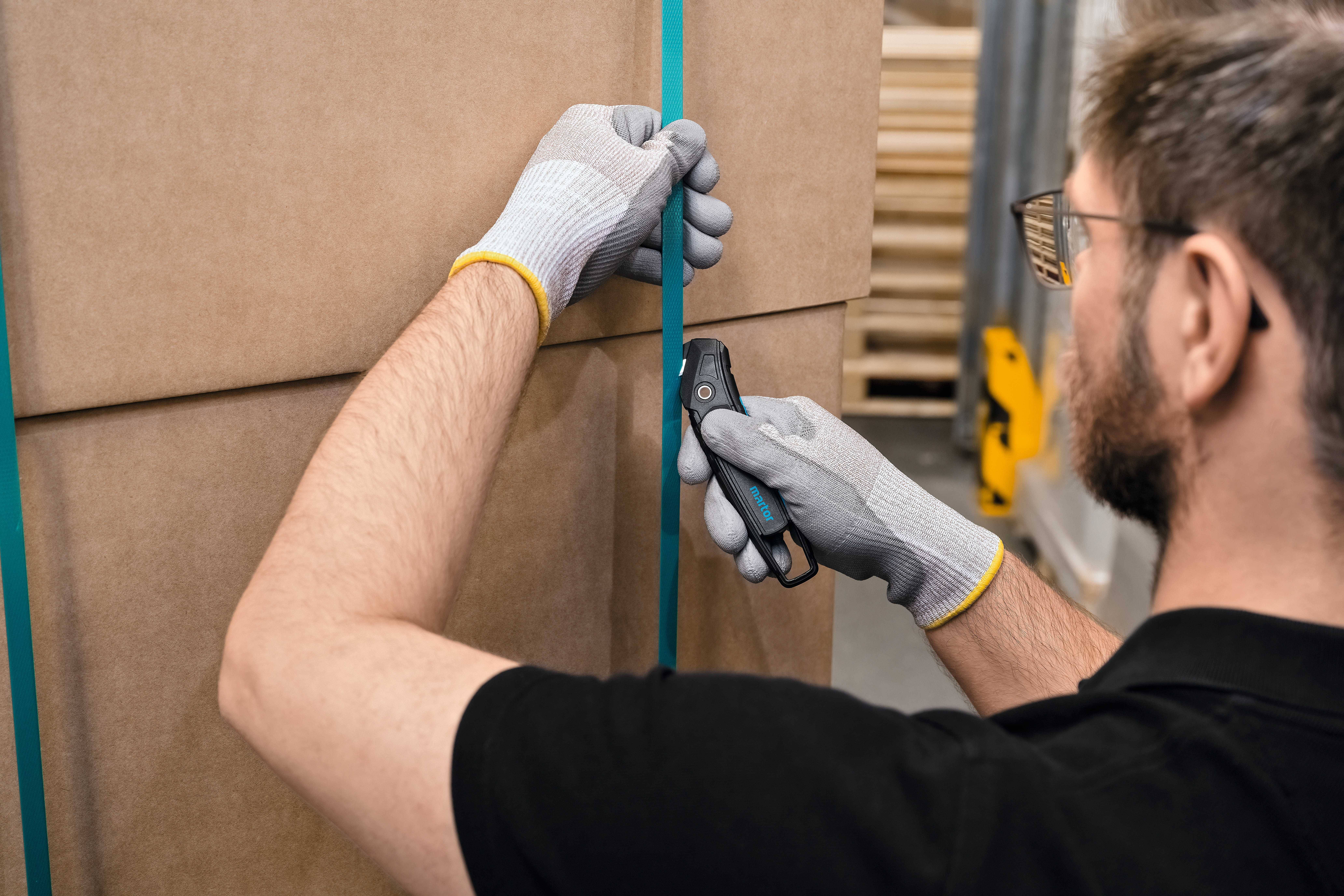 Een persoon draagt handschoenen en snijdt met een gereedschap het pakketband van een kartonnen doos.