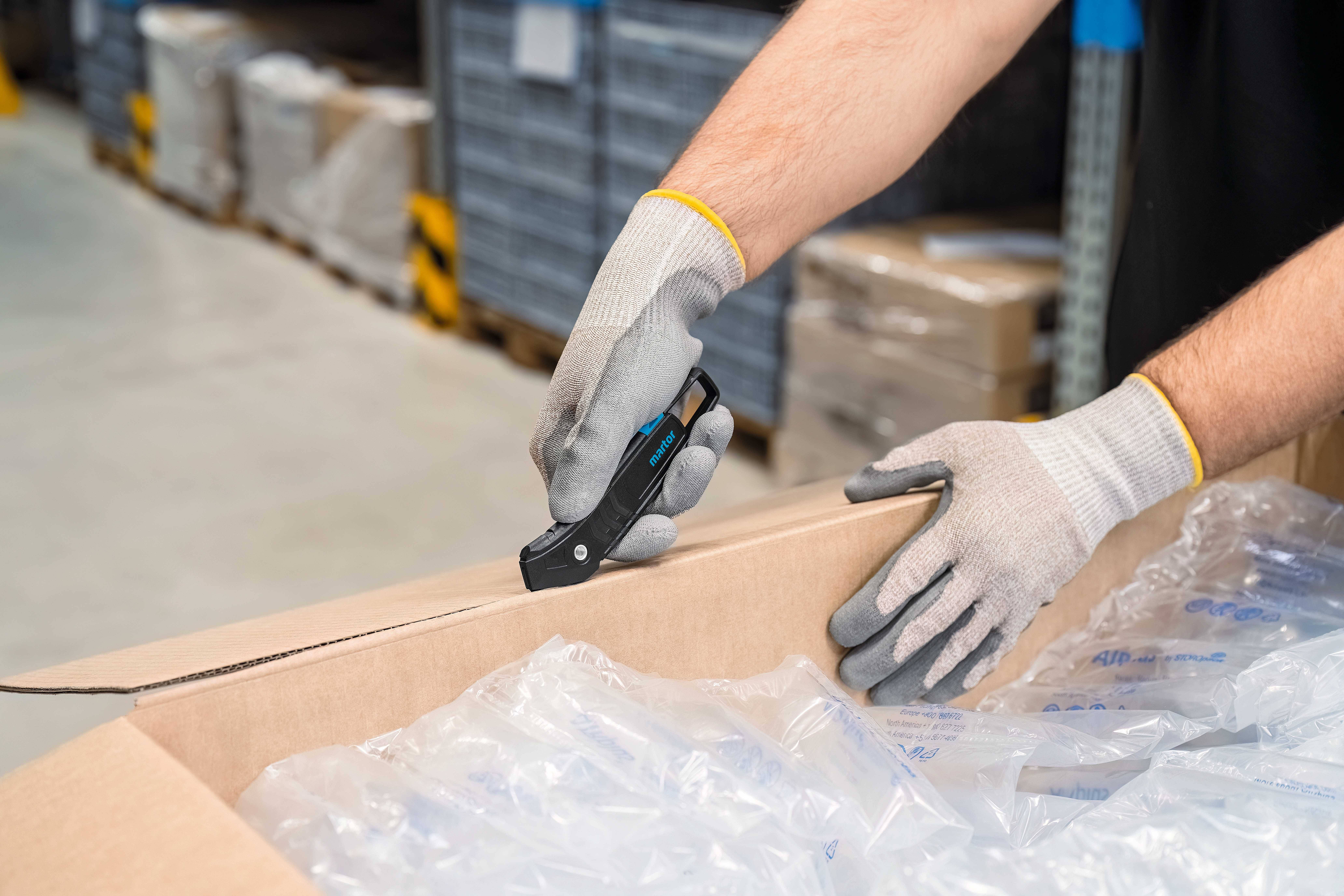 Persoon opent een karton in een magazijnhal met een veiligheidsmes. De doos is gevuld met luchtkussenfolie.