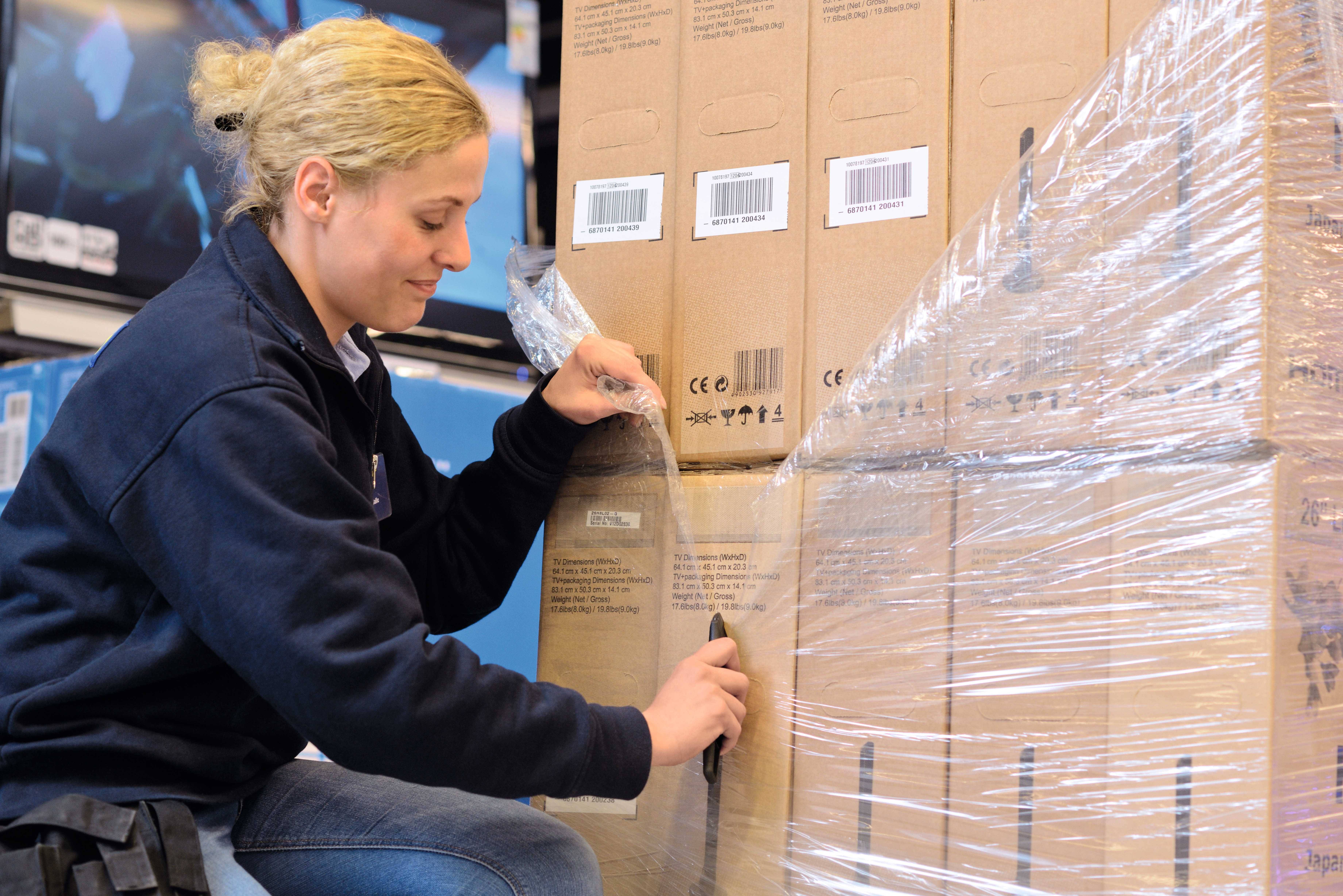 Een persoon opent met een mes een pallet met dozen in een magazijn.