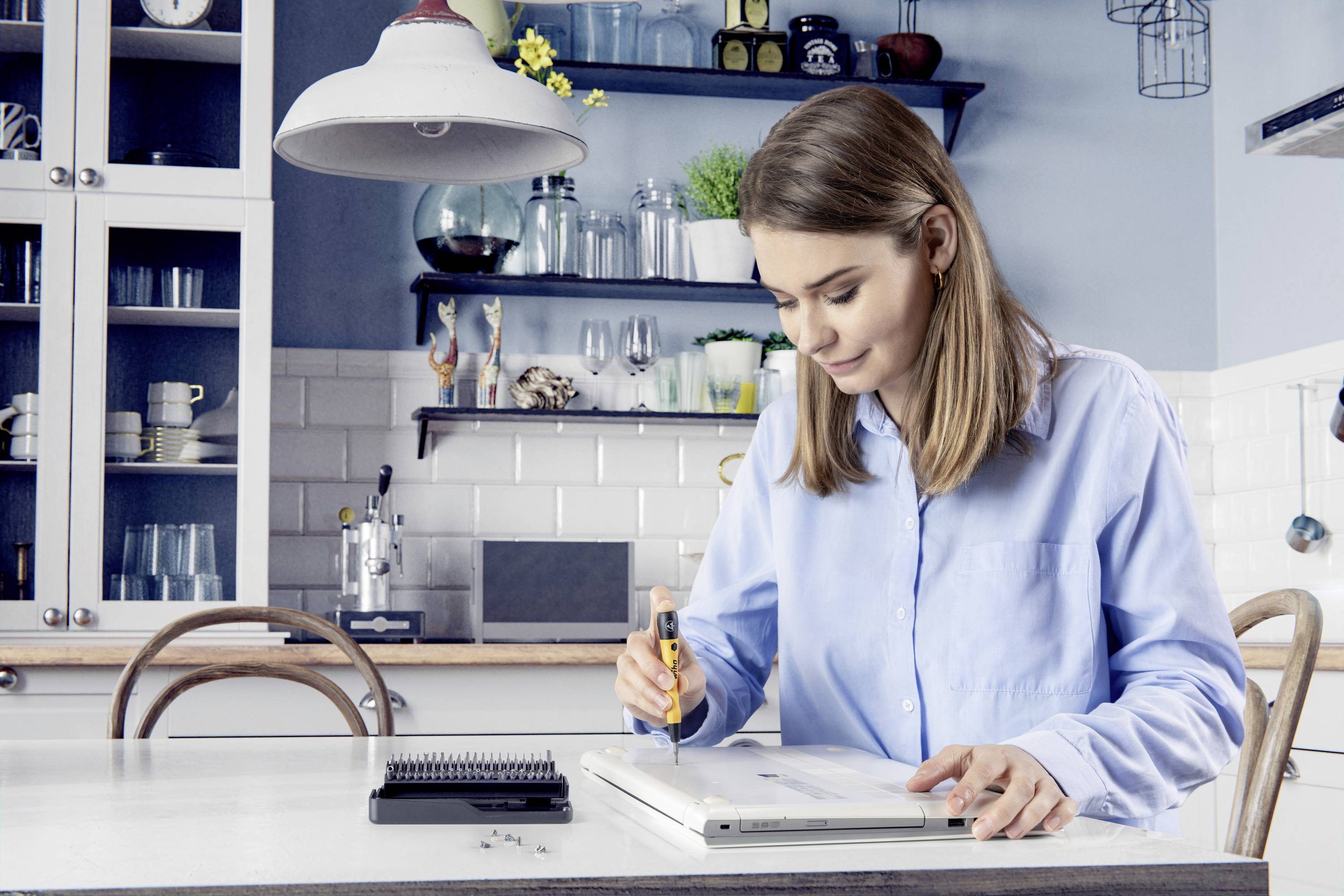 Een vrouw zit in de keuken en repareert een laptop met een schroevendraaier. Op tafel liggen gereedschap en schroeven.