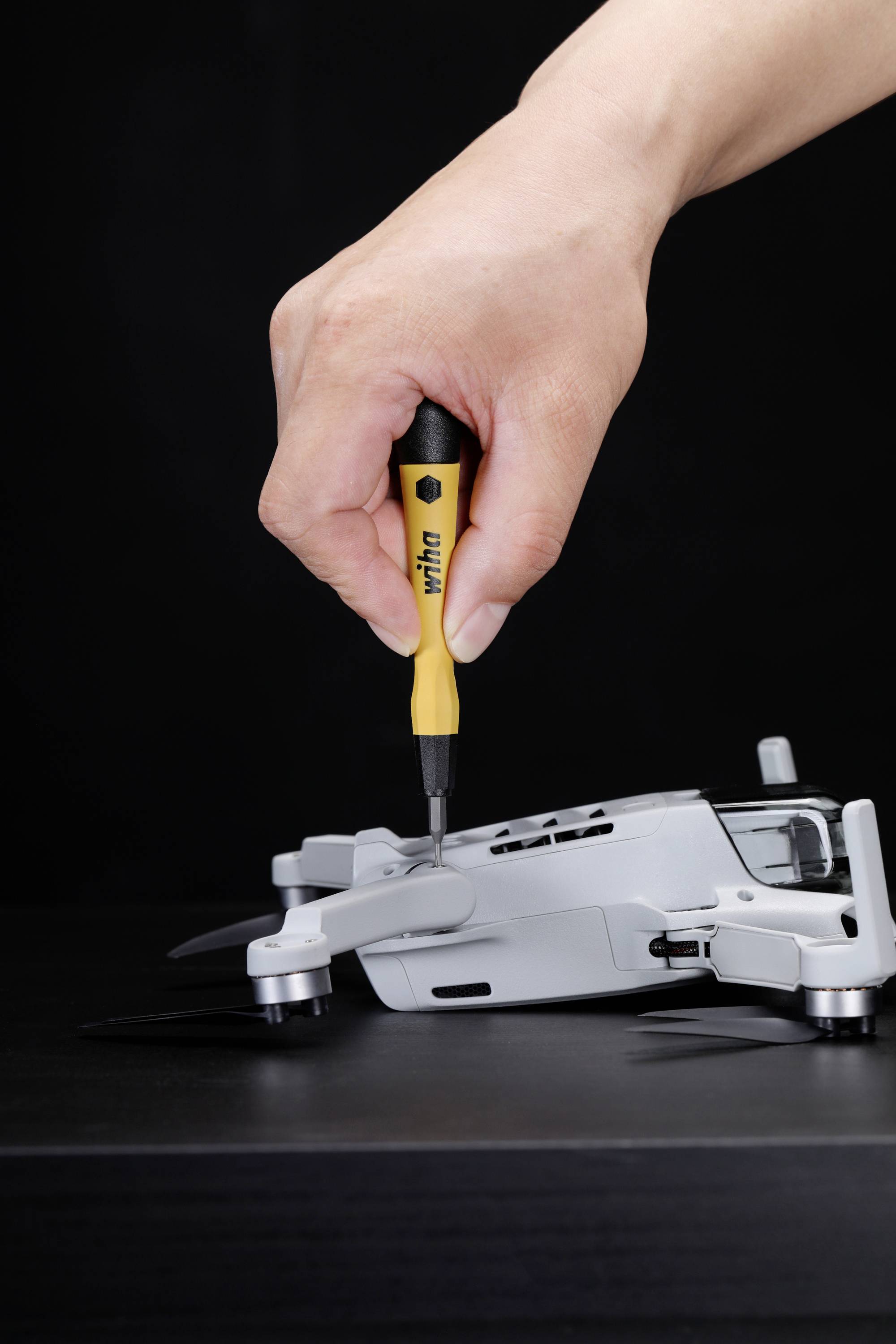 Een hand repareert een drone met een schroevendraaier. De drone ligt plat op een donker oppervlak.