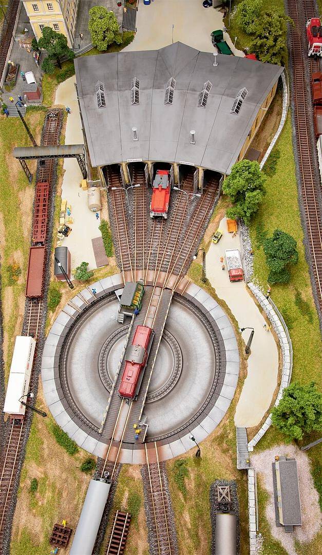 Een modeltreinendepot met locomotieven op sporen, een draaischijf in het midden, omgeven door bomen en andere modelgebouwen.