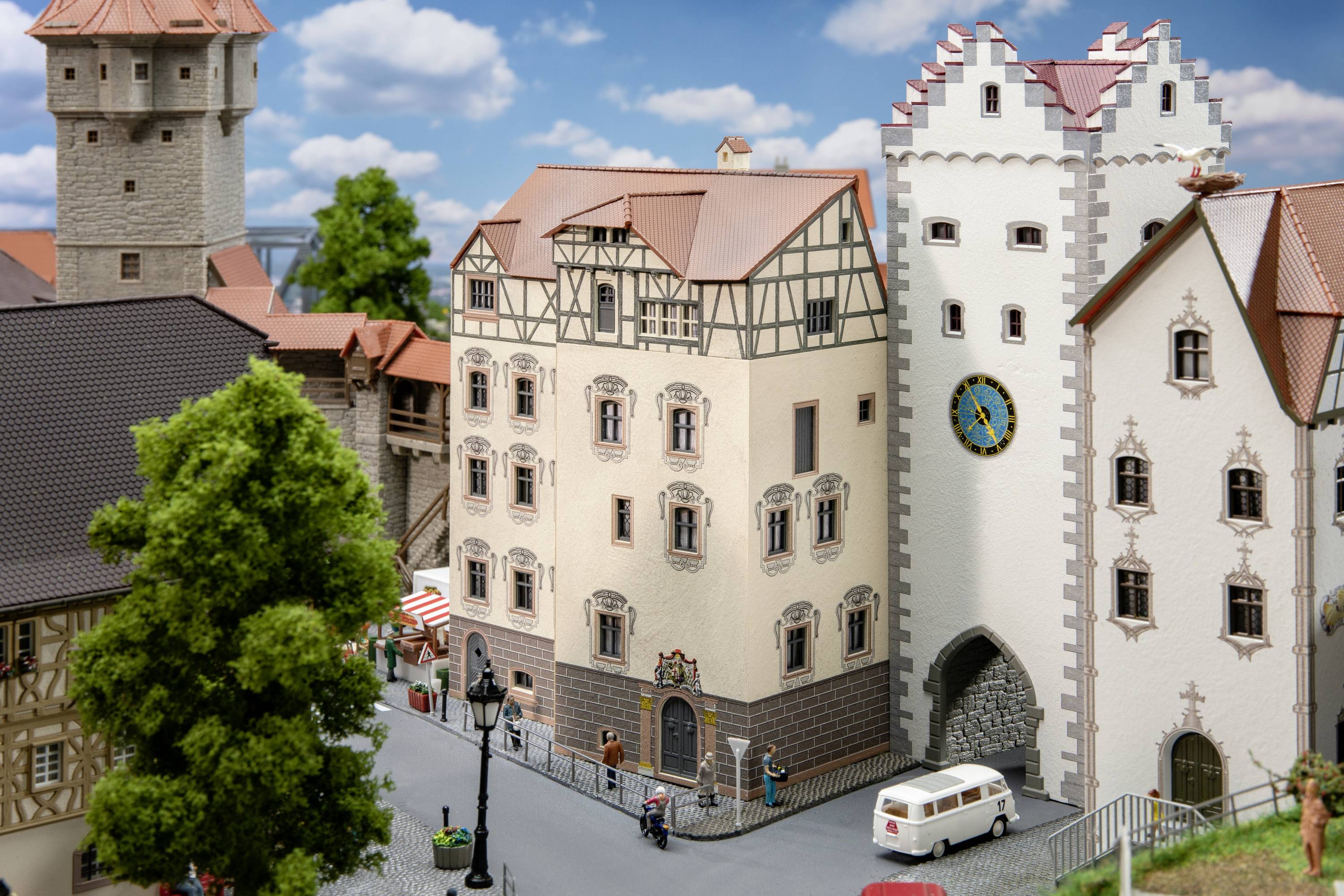 Miniatuurstad met historische gebouwen, een poort met een klok, een auto en mensen op een geplaveid straatje, bomen op de achtergrond.