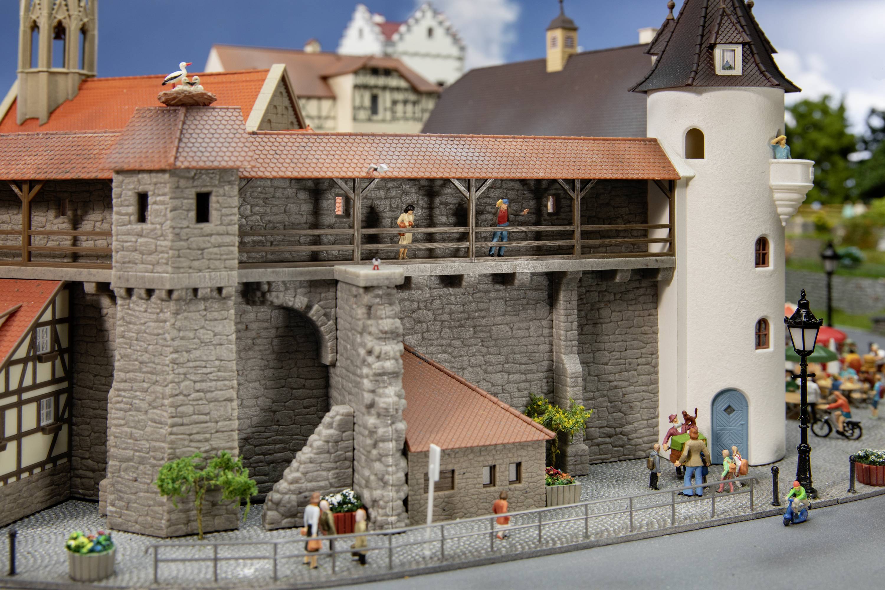 Miniatuurmodel van een middeleeuwse stadspoort met mensen op de doorgang en in de omgeving. Gebouwen op de achtergrond, blauwe lucht.