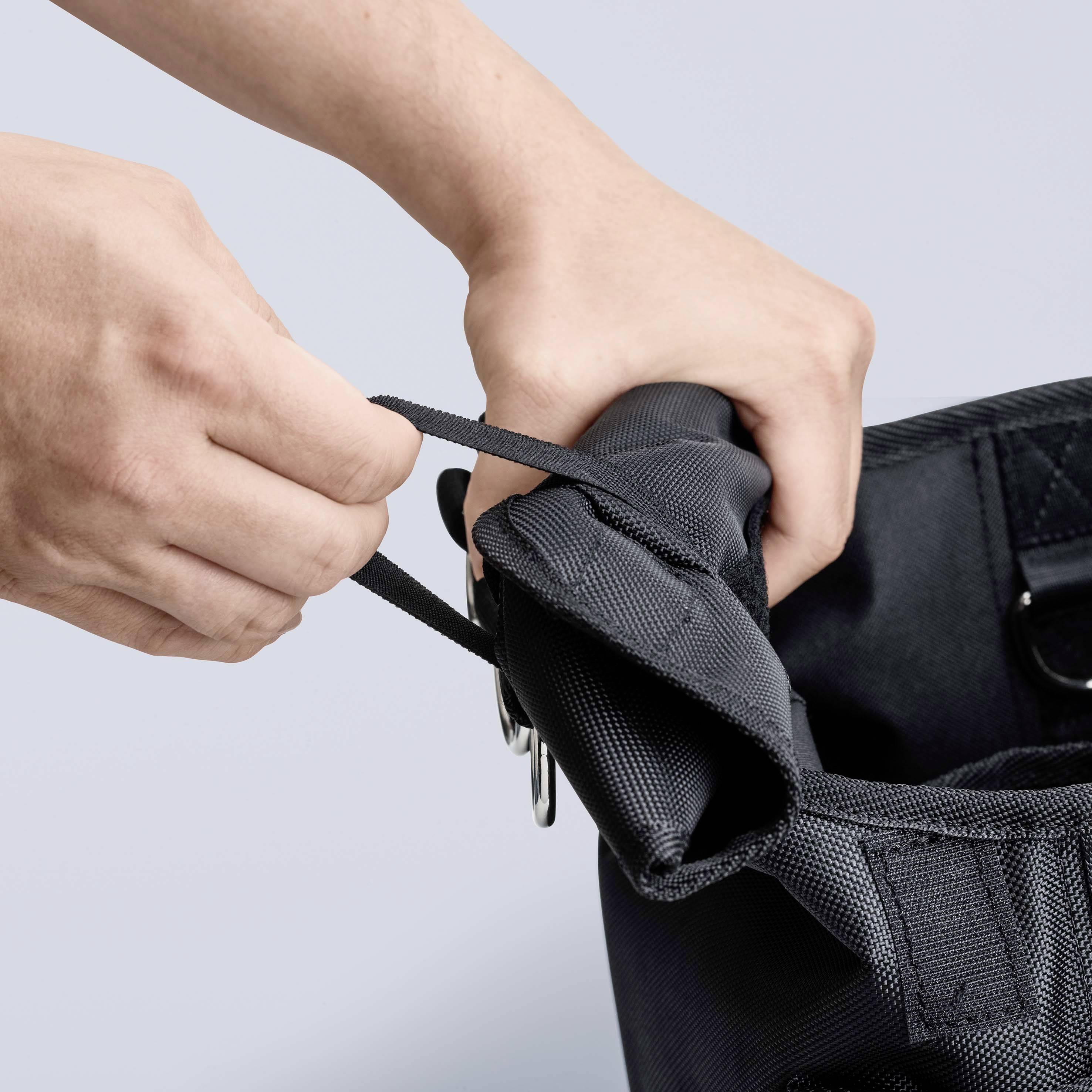 Een persoon trekt aan een zwarte rugzakband om deze bij te stellen. Handen en rugzak zijn zichtbaar, de achtergrond is neutraal.