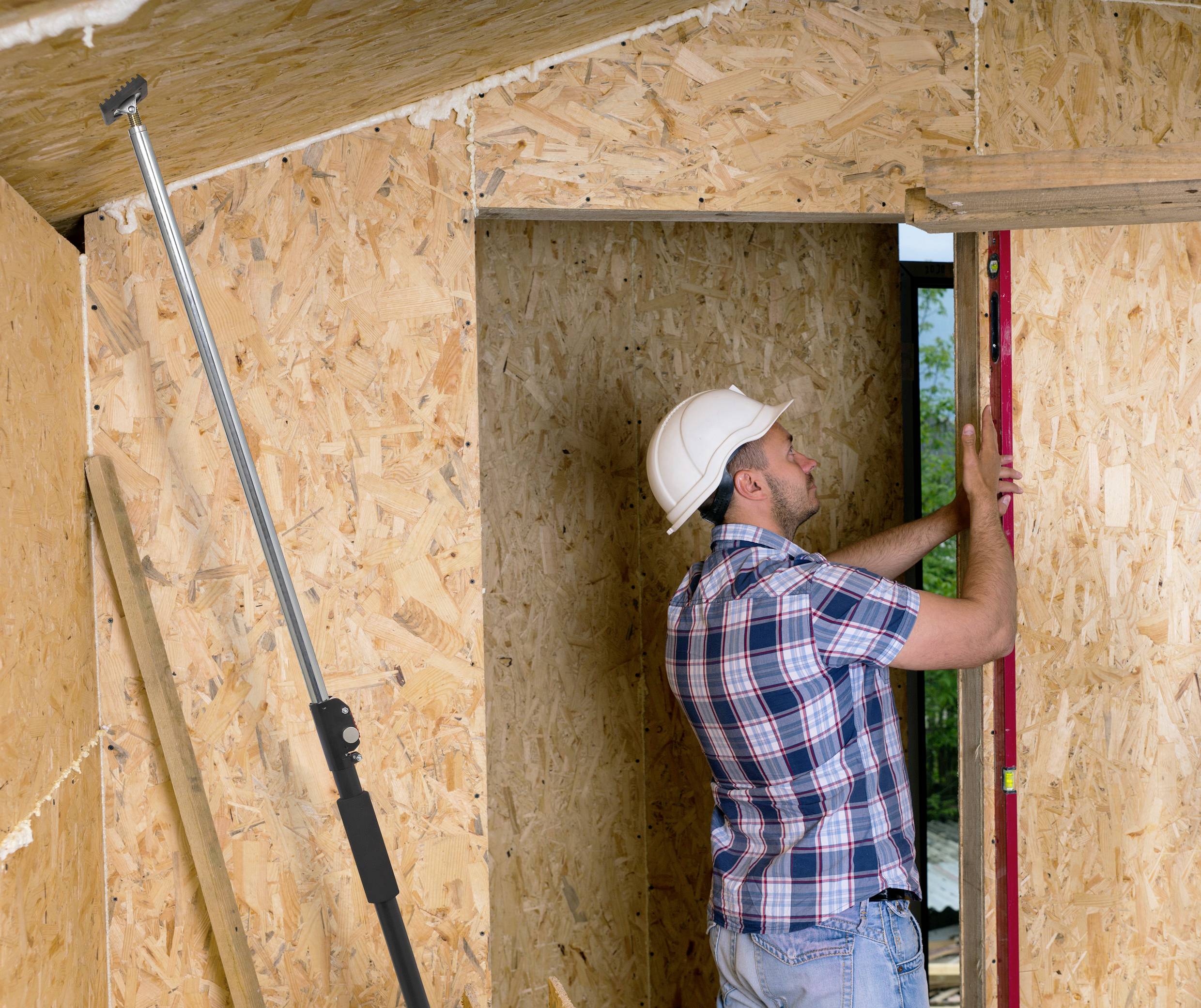 Een bouwvakker in een geruit overhemd en helm richt een houten wand uit met een waterpas. Hij staat in een gedeeltelijk afgewerkte ruimte.
