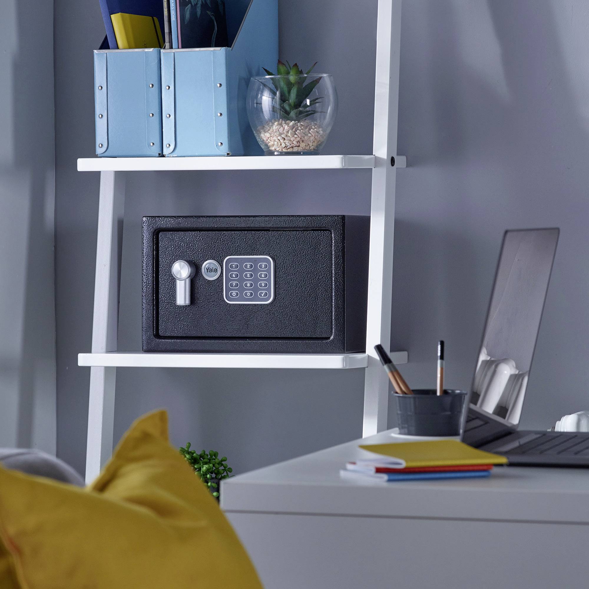 Een wit rek met een kluis, boeken en een glas met een plant erop. Naast het rek ligt een laptop en een notitieboek op een bureau.