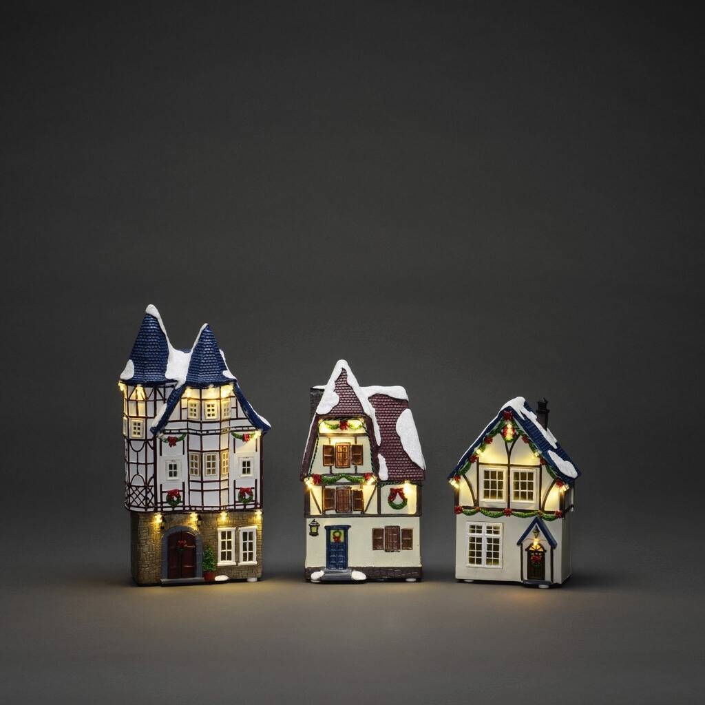 Drie verlichte modellhuizen met verschillende architecturale stijlen op een donkere achtergrond, die een winterse scène verbeelden.