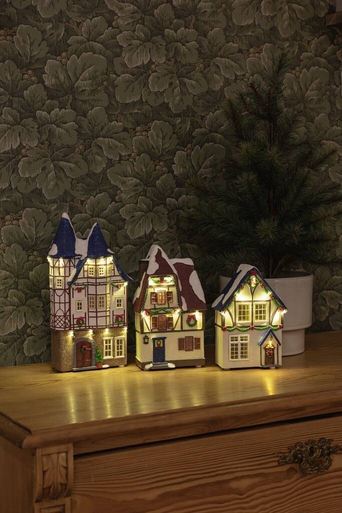 Drie verlichte miniatuurhuisjes op een houten tafel voor een gedessineerd behang, decoratief gerangschikt in een gezellige omgeving.