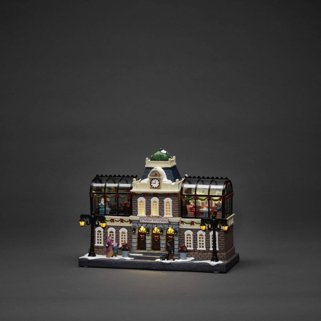 Een verlicht model van een station in de winter, versierd met sneeuwval, figuurtjes en een klokkentoren, tegen een donkere achtergrond.