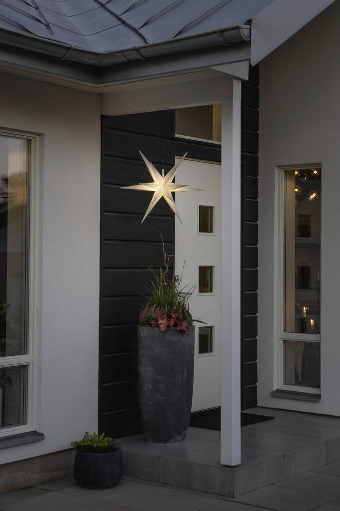 Een verlichte ster hangt aan de buitenmuur van een modern huis. Ernaast staat een hoge plantenbak met bloemen.
