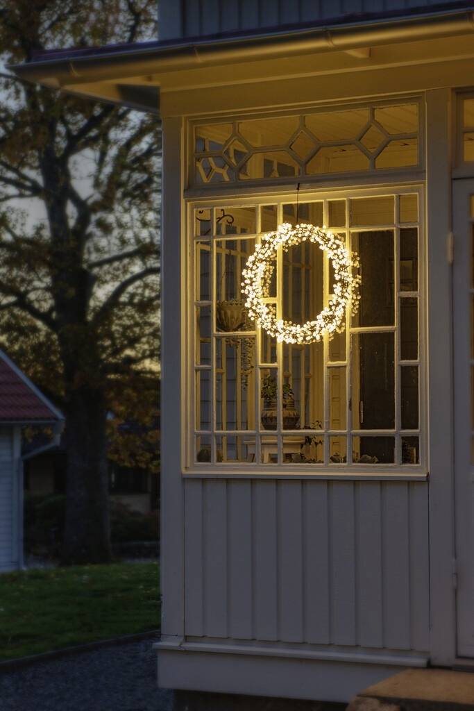 Een verlichte kerstkrans hangt in het raam van een huis bij schemering, stralend in contrast met de donkere omgeving.