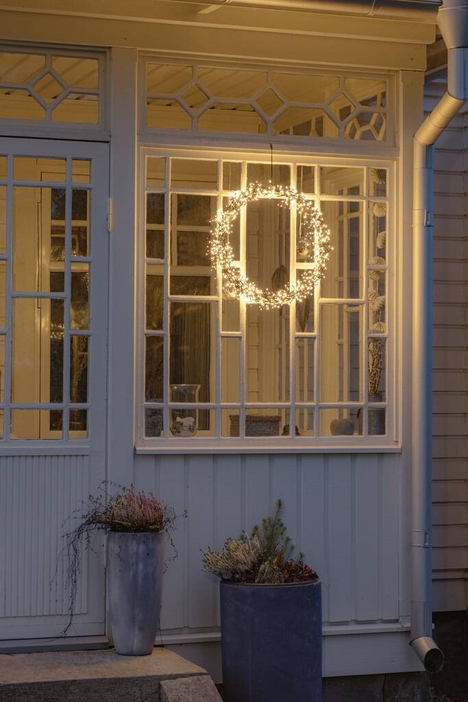 Een verlichte krans hangt aan de binnenkant van een raam in een huis met decoratieve lijsten. Buiten staan twee bloempotten op de veranda.