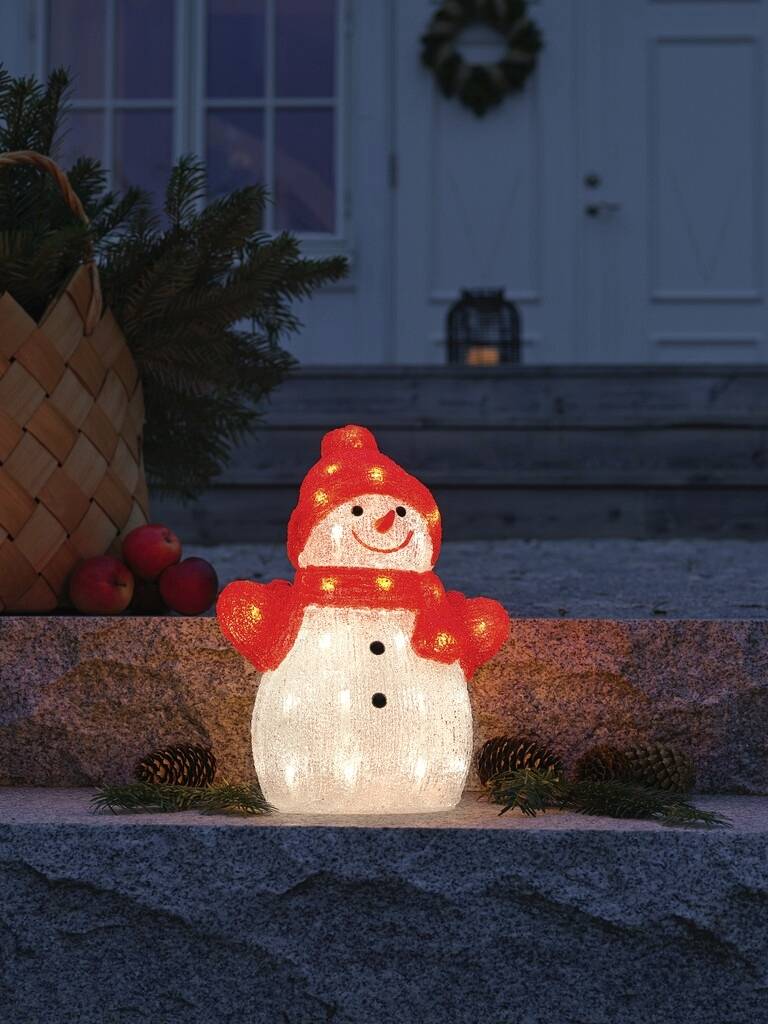 Een stralende sneeuwpop-decoratie met een rode hoed en sjaal staat op een trap naast dennenappels en een mand met appels.