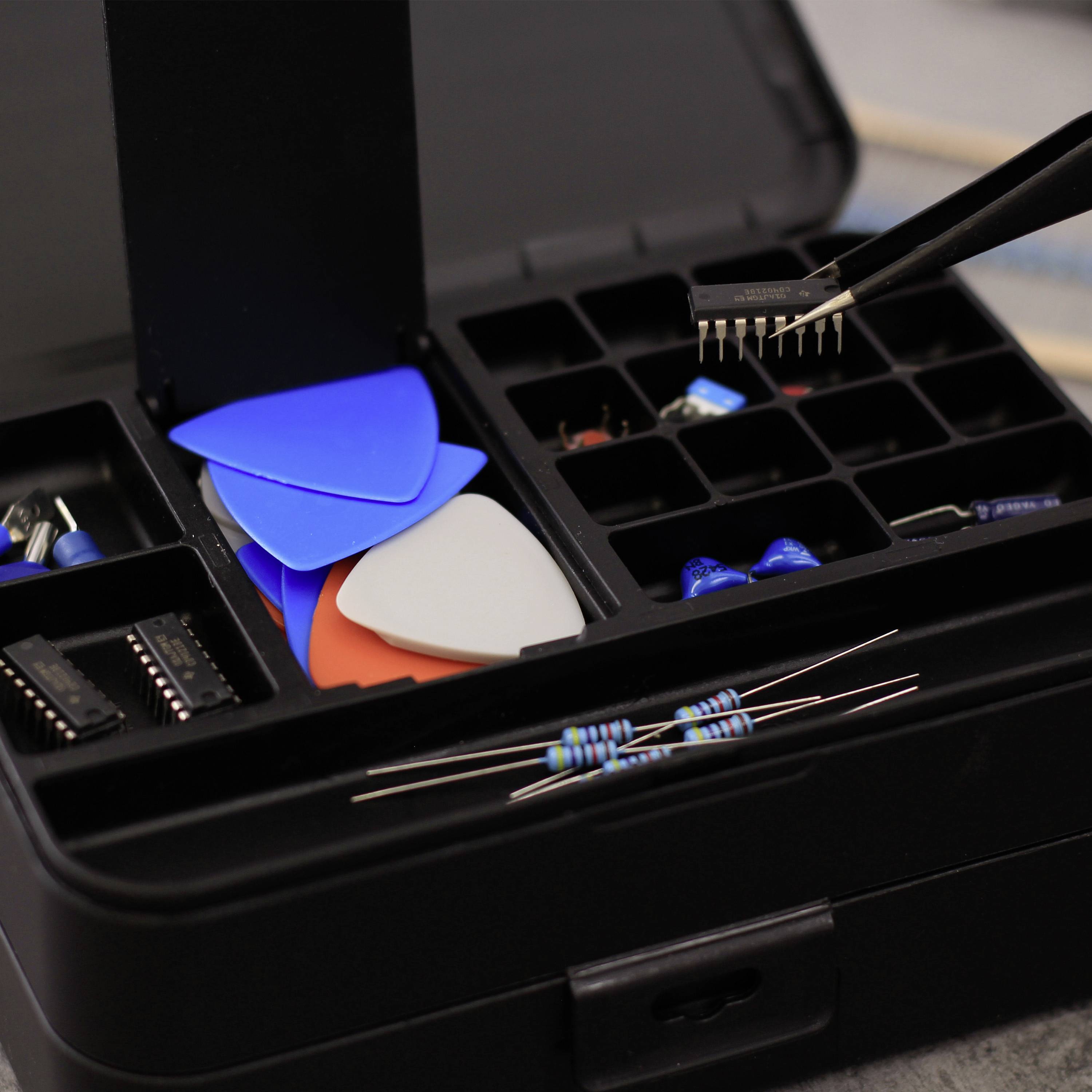 Gereedschapskist met elektronische componenten: weerstanden, chips, plectrums en een pincet dat kleine pinnen vasthoudt.