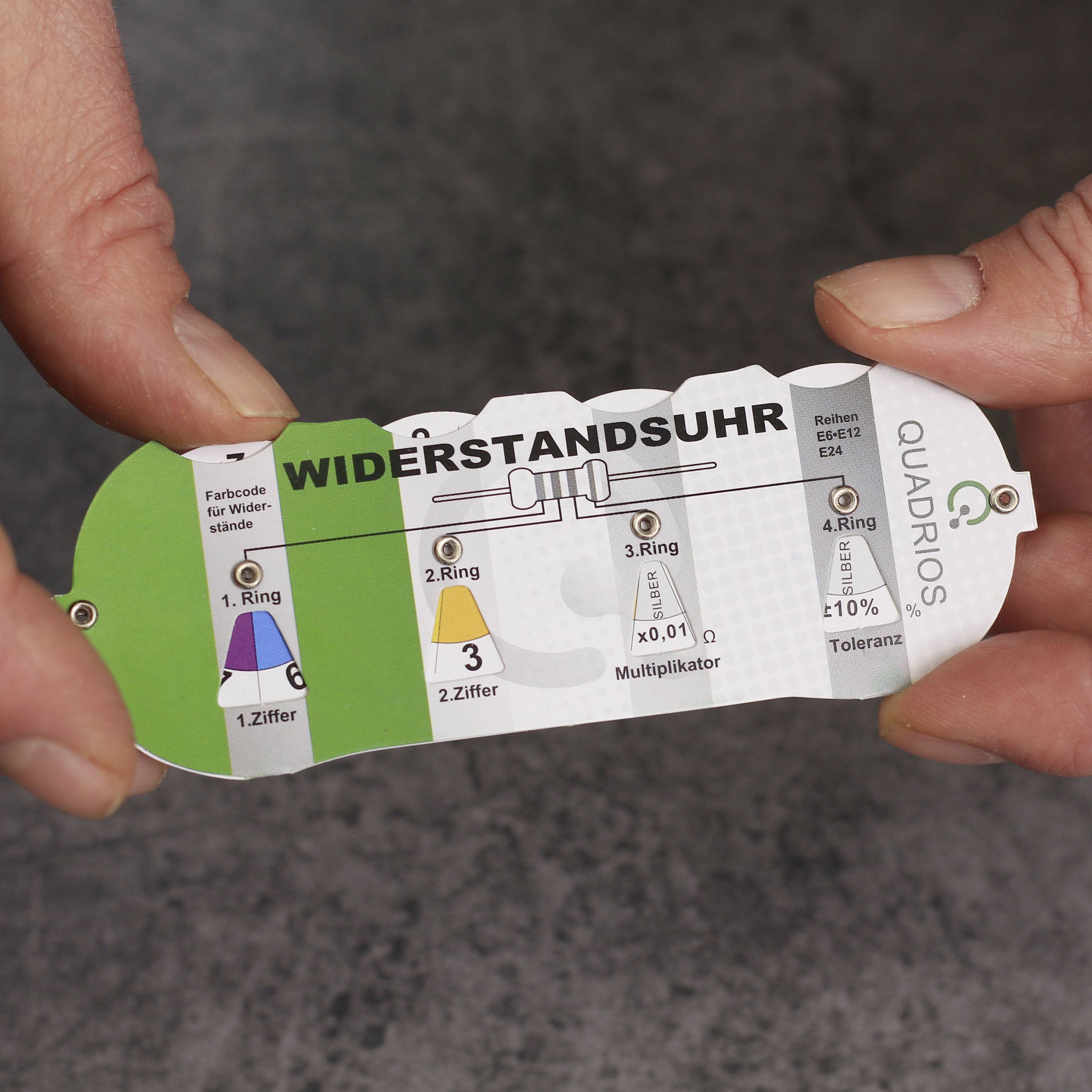 Twee handen houden een weerstandsmeetkaart vast, die laat zien hoe men de waarde van een elektrische weerstand met kleurringen kan aflezen.
