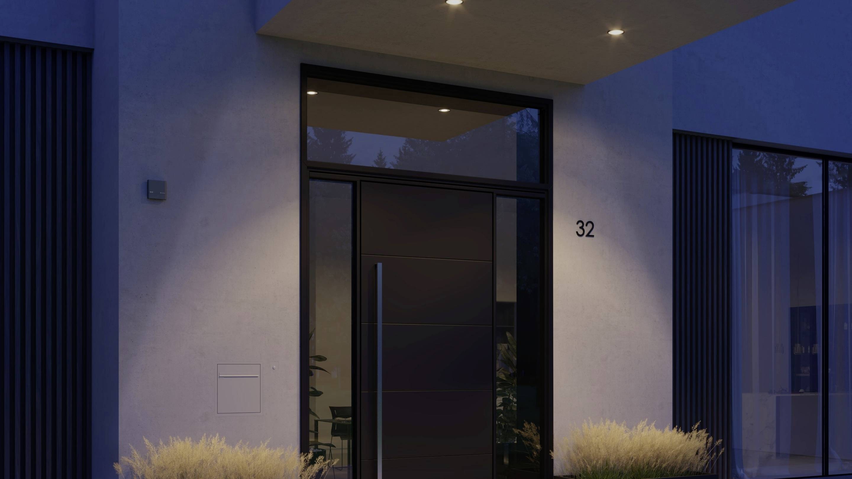Moderne voordeur van een huis bij het vallen van de avond, verlicht door buitenverlichting, met huisnummer 32. Grasstrook op de voorgrond.