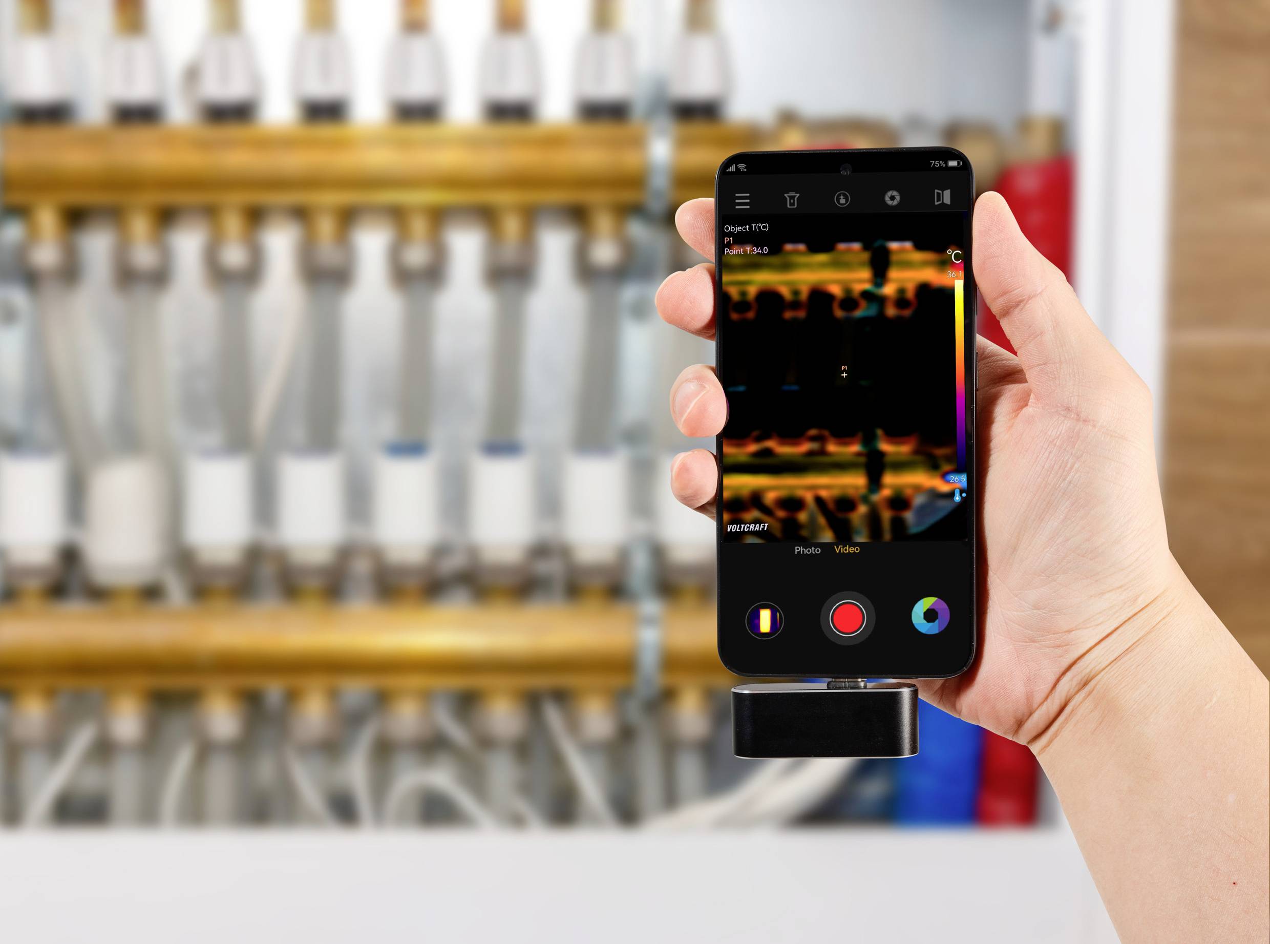 Een hand houdt een smartphone vast waarop een warmtebeeldopname van een waterleiding wordt weergegeven. Op de achtergrond zijn onscherpe buisleidingen zichtbaar.