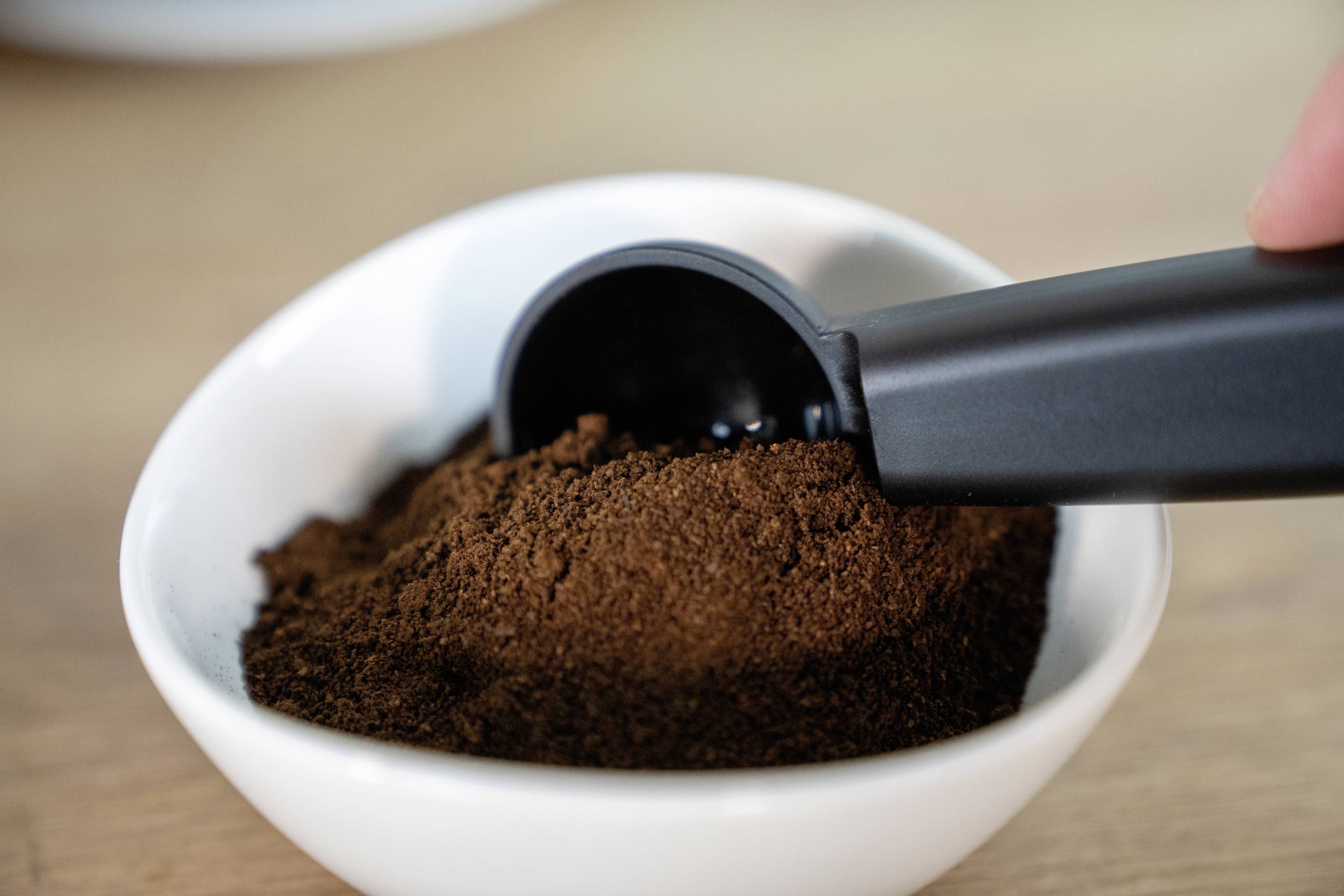 Een schaal met gemalen koffie, waar een zwarte lepel in steekt, op een houten tafel.