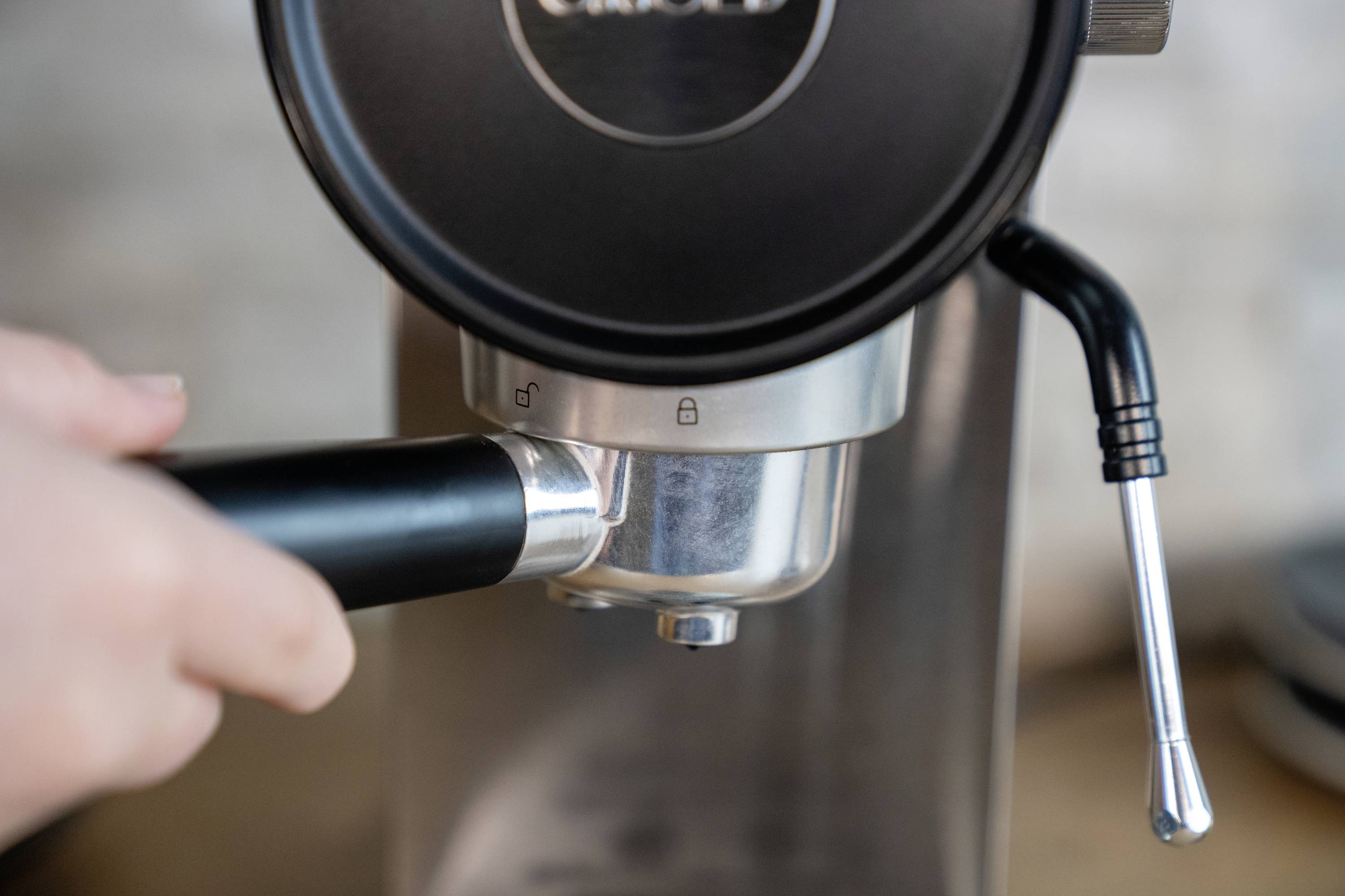 Een hand houdt de filterhouder van een zilveren espressomachine vast. Een stoomtuit is zichtbaar aan de rechterkant.