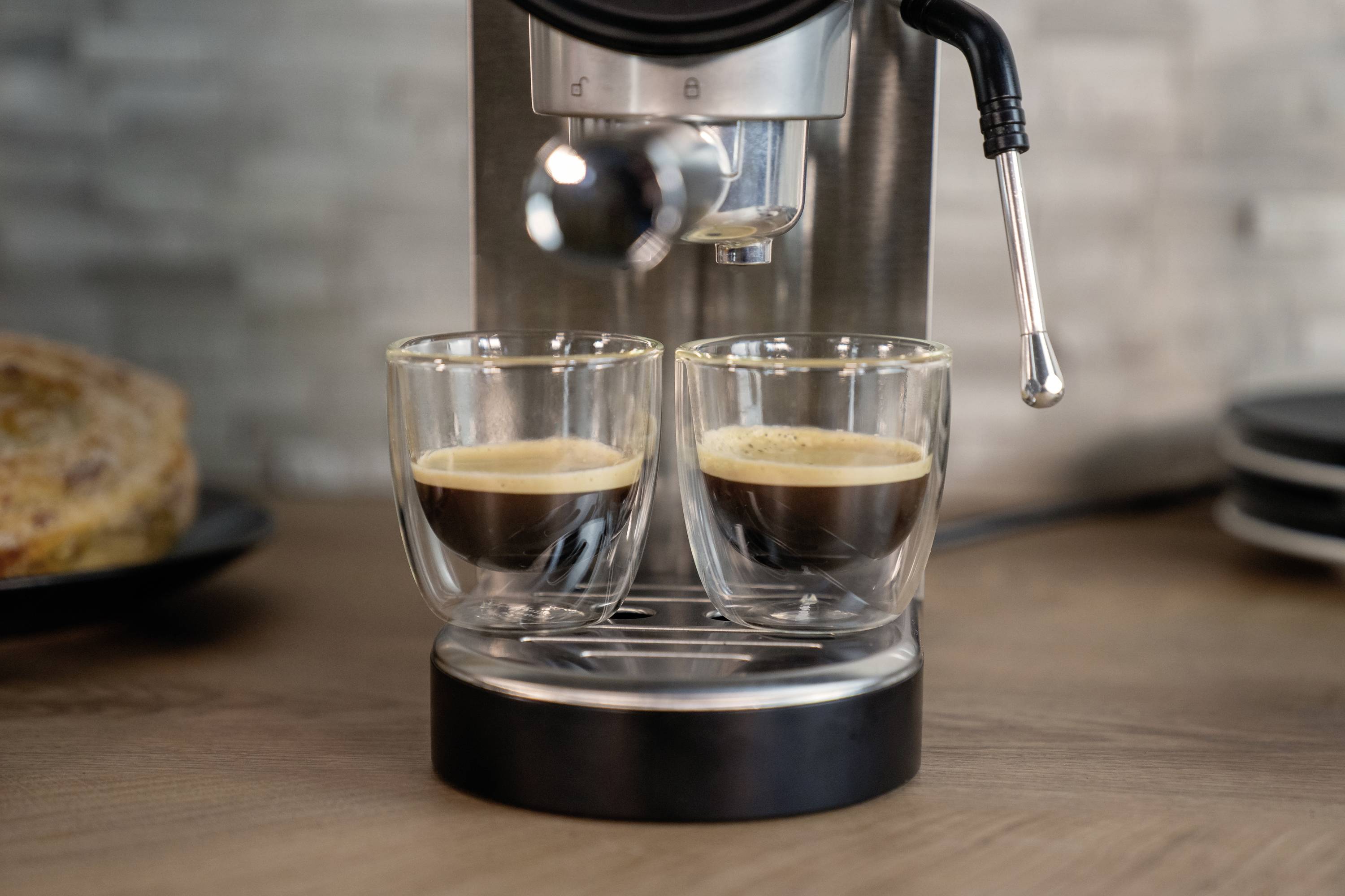 Twee espressoglazen onder een koffiemachine die koffie inschenkt. Op de achtergrond een bord met brood.