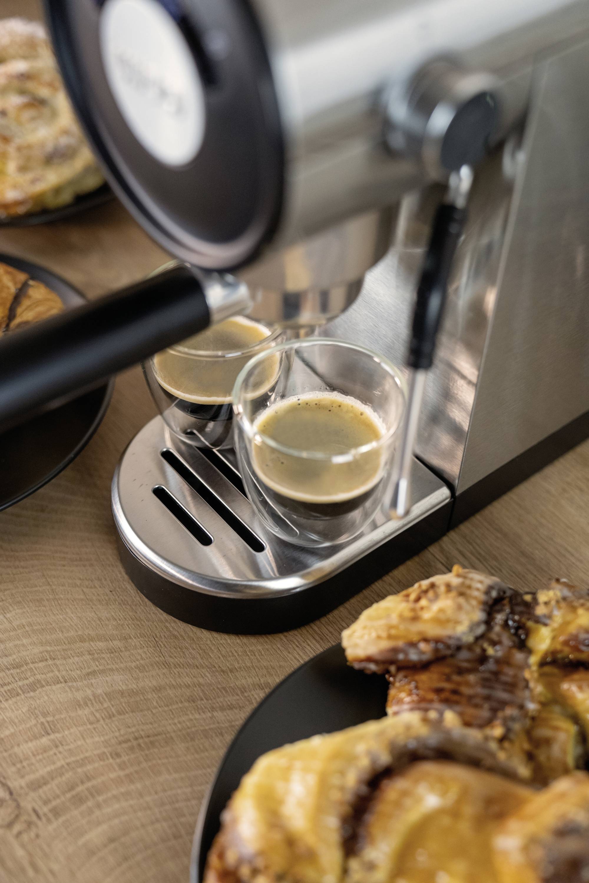Espressomachine schenkt verse espresso in twee glazen. Ernaast liggen verschillende gebakjes op een houten tafel.