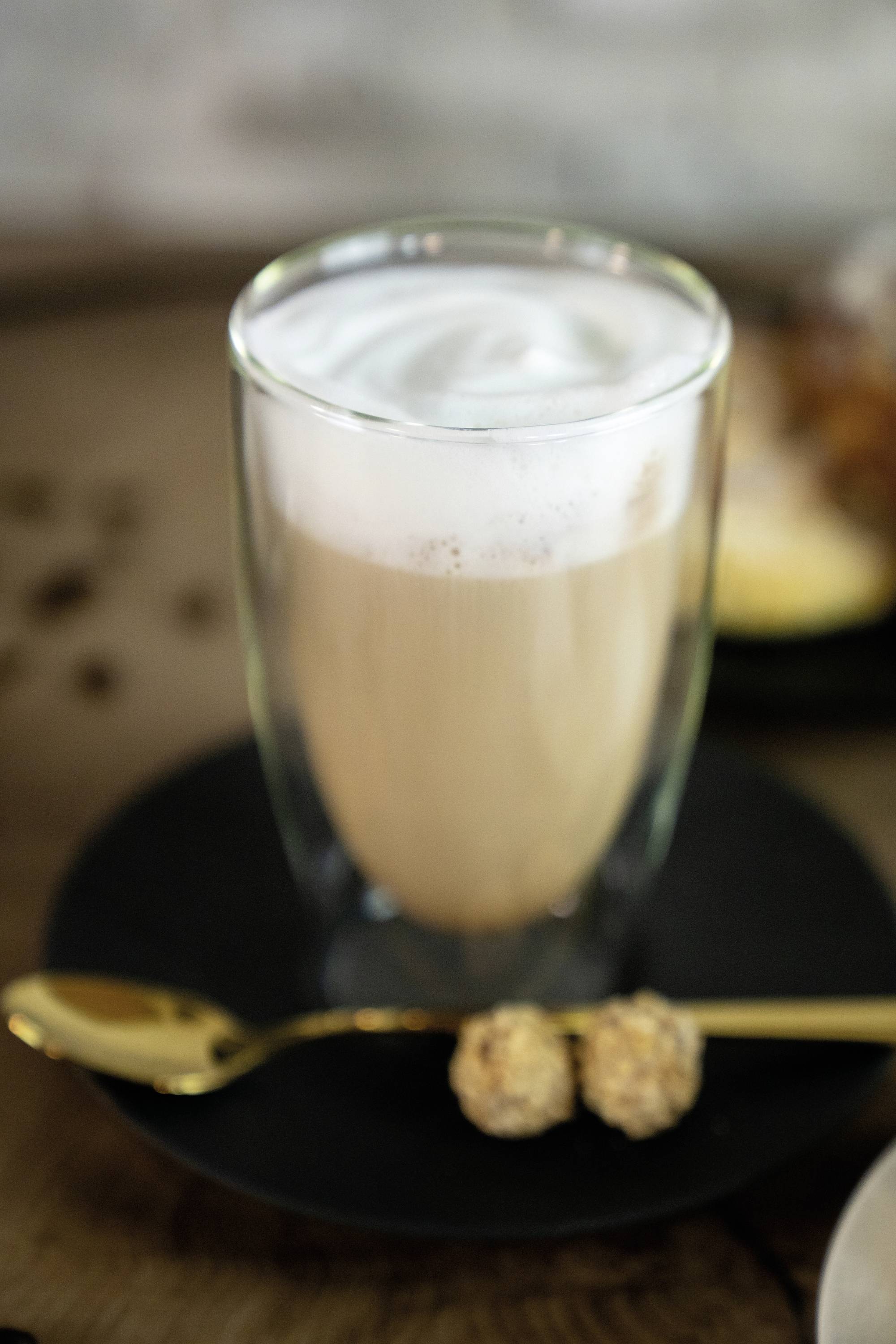 Een glas koffiedrank met romige melkschuim, geserveerd op een bord met twee pralines naast op een gouden lepel.