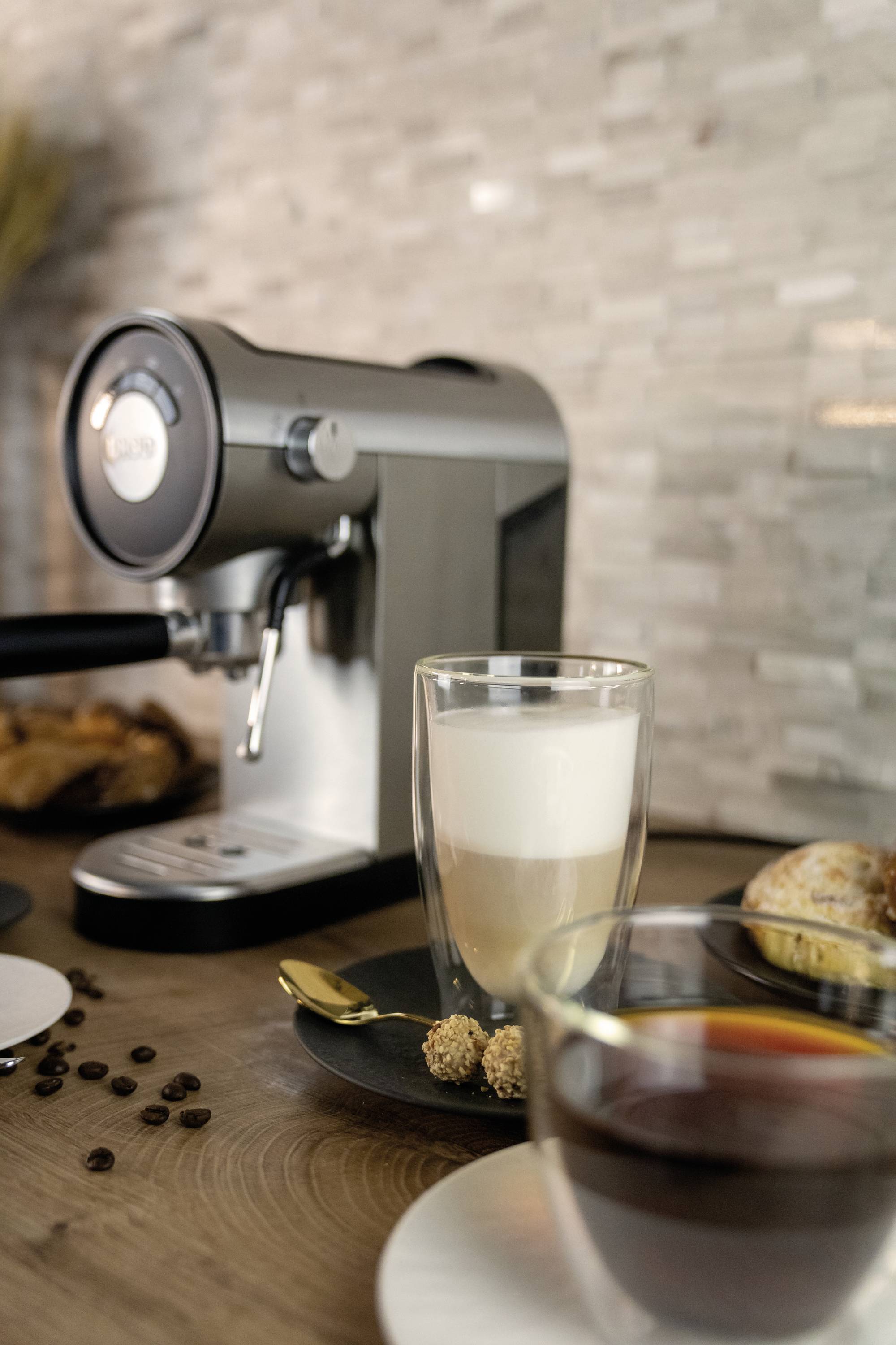 'Koffiemachine met een kopje cappuccino op de voorgrond, omringd door croissants en koffiebonen op een houten tafel.'