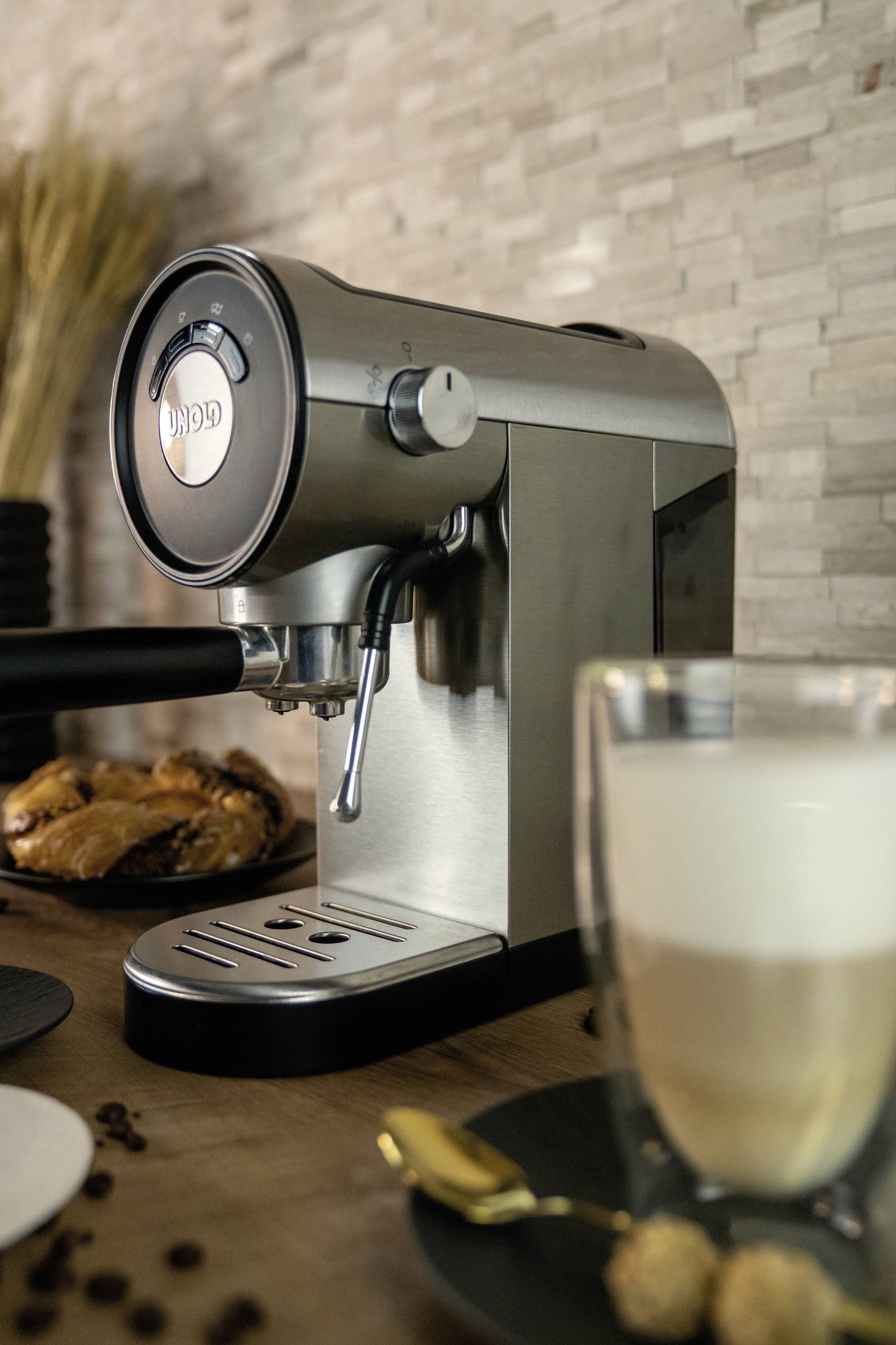Zilveren espressomachine met een latte macchiato glas en gebak op een houten tafel, met een gestructureerde wandbekleding op de achtergrond.