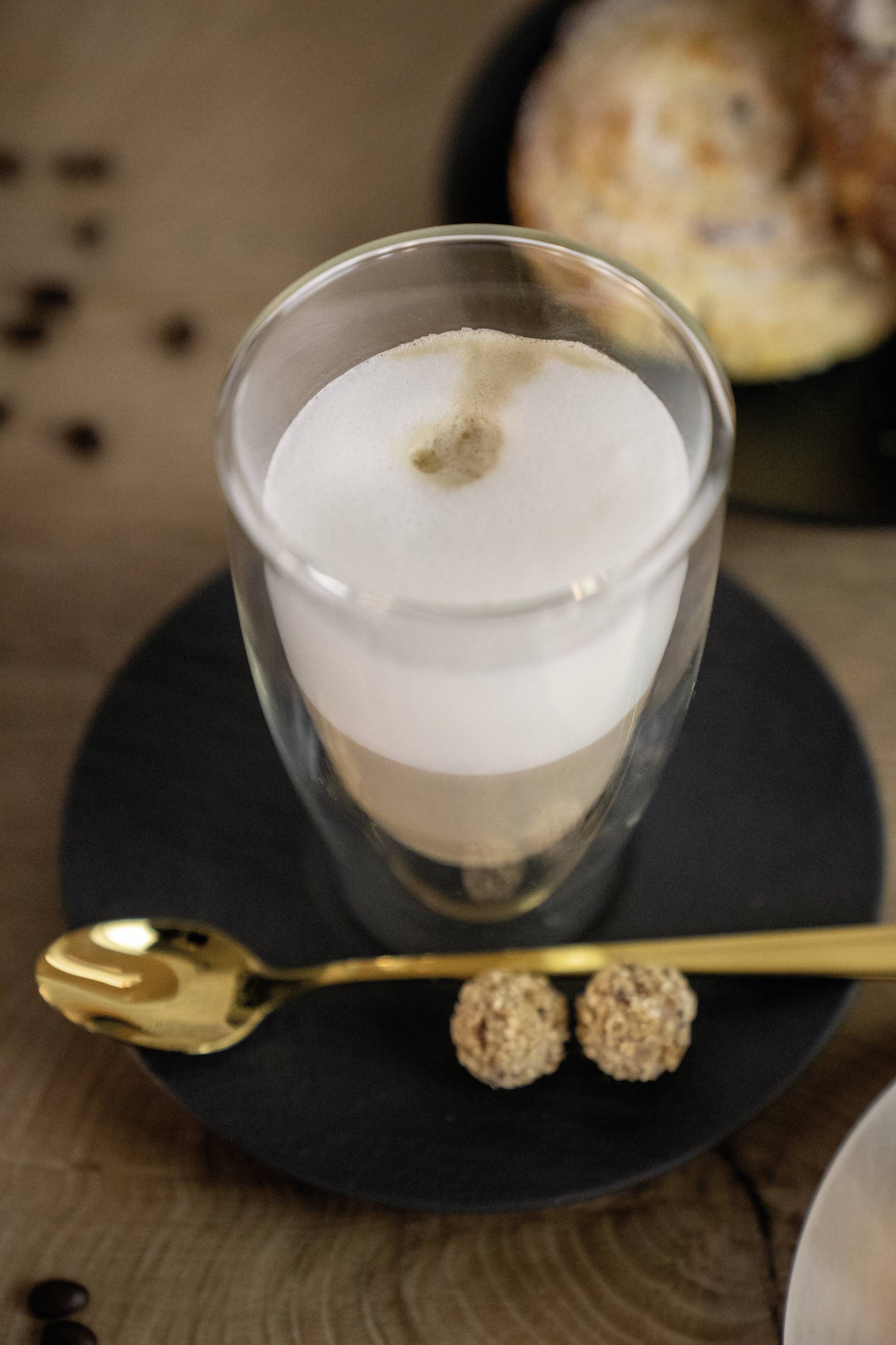 Een glas Latte Macchiato op een zwarte schotel met een gouden lepel en twee kleine koekjes. Op de achtergrond een gebak.
