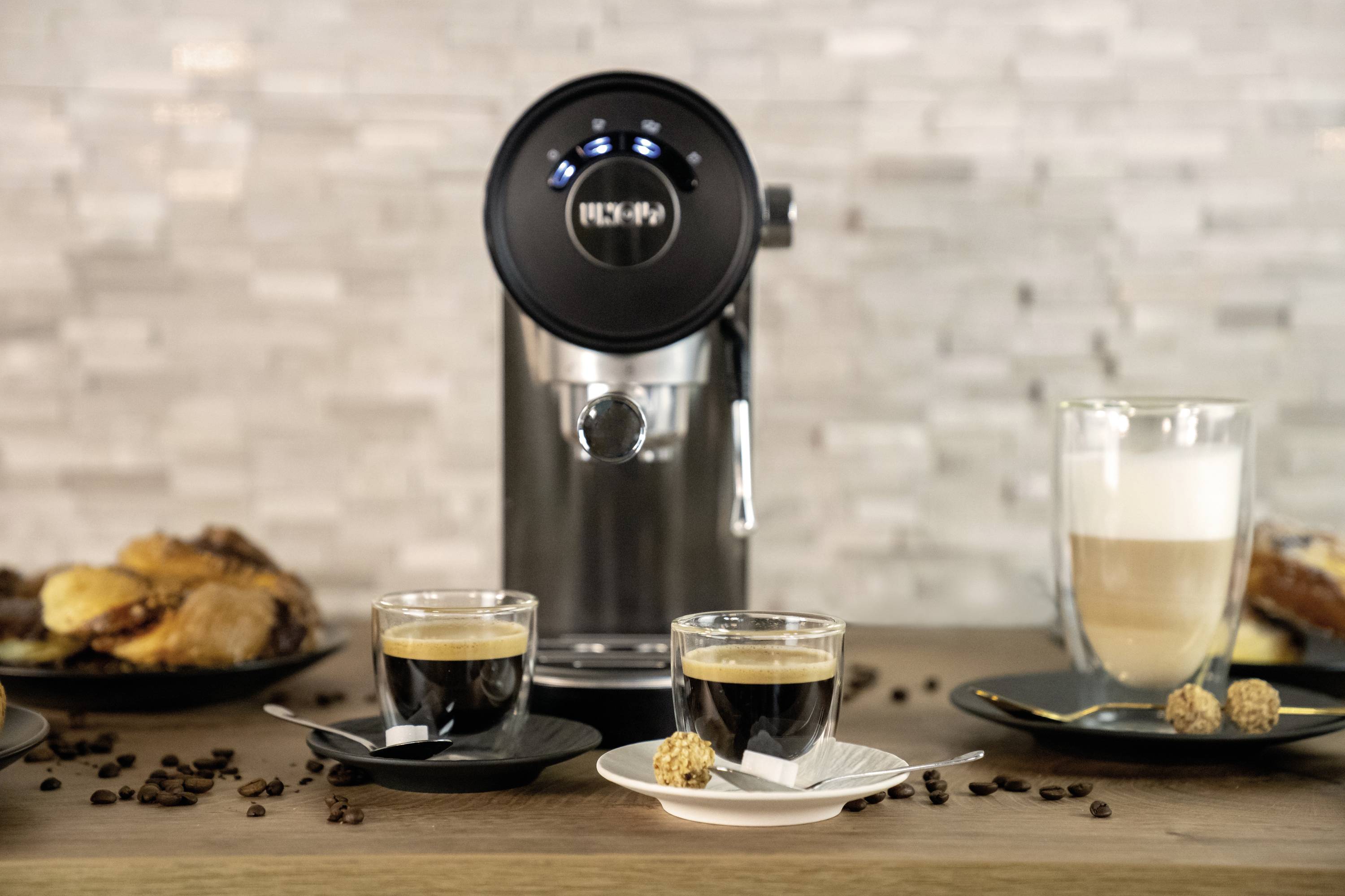 Koffiemachine met twee espresso's in glazen, naast een latte macchiato, op de achtergrond croissants en koffiebonen op een tafel.