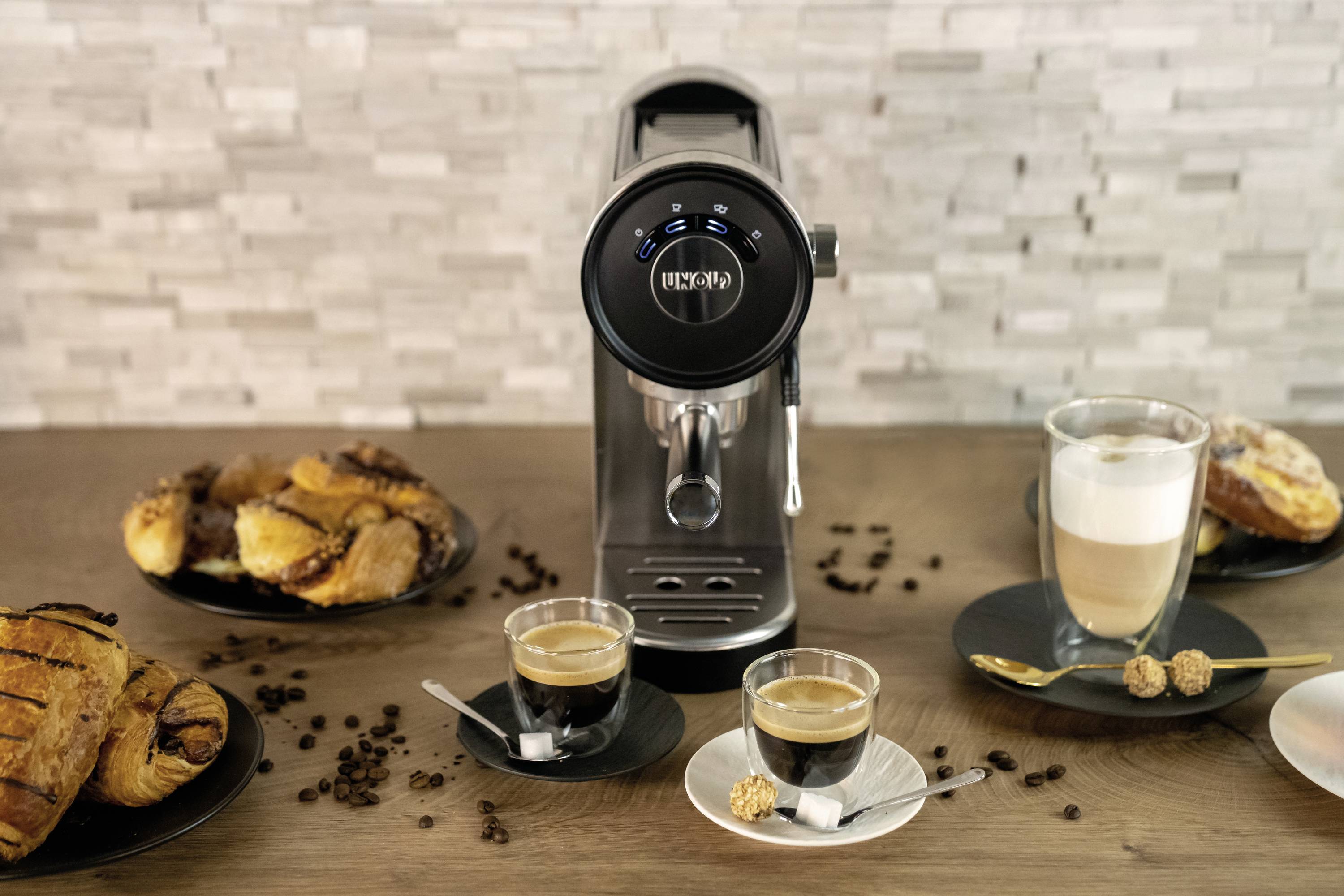 'Een espressomachine met twee gevulde kopjes koffie op een tafel, omringd door gebak en koffiebonen, voor een mozaïekwand.'