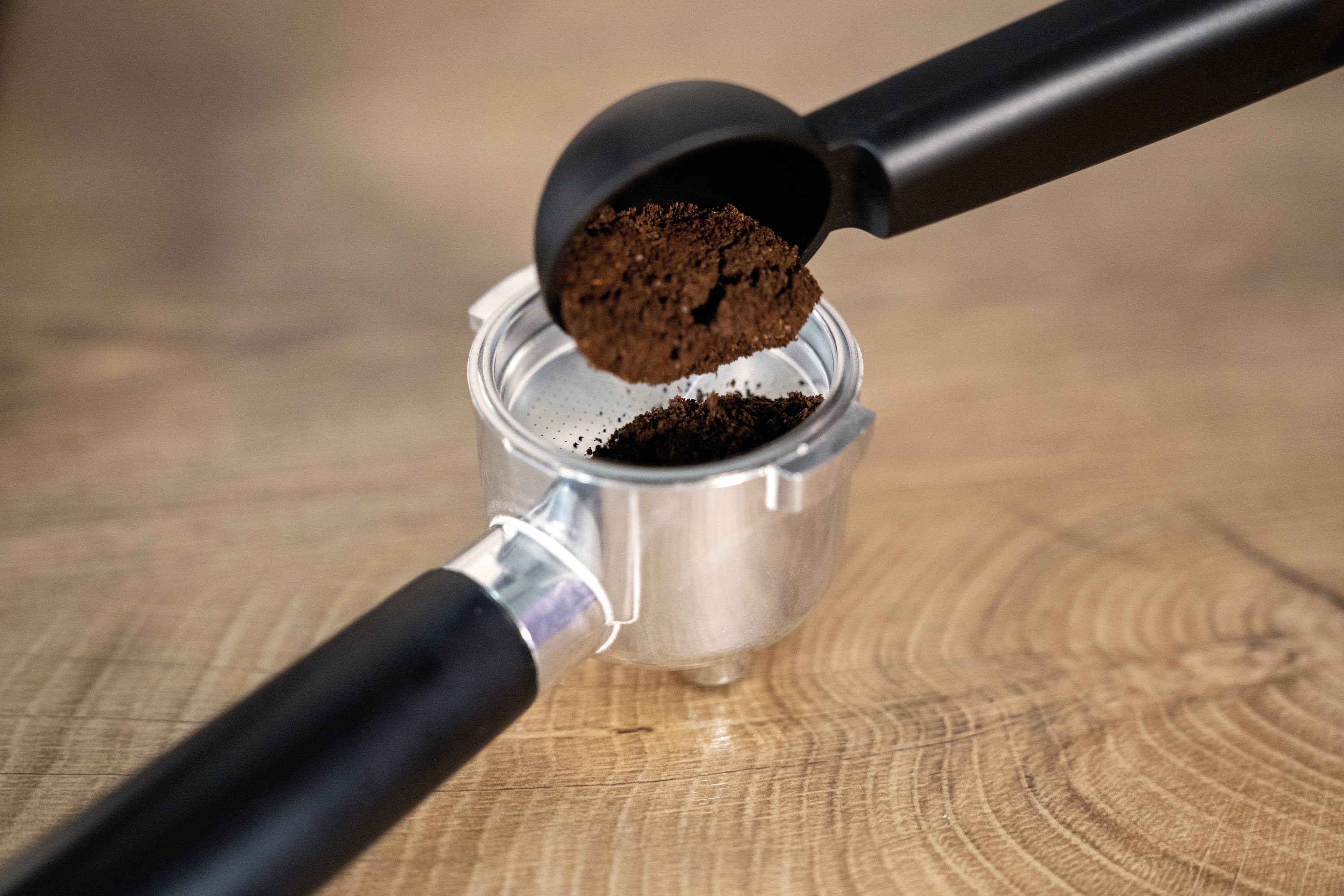 Een lepel gemalen koffie wordt in een filterhouder boven een houten oppervlak gedaan, klaar voor het bereiden van espresso.