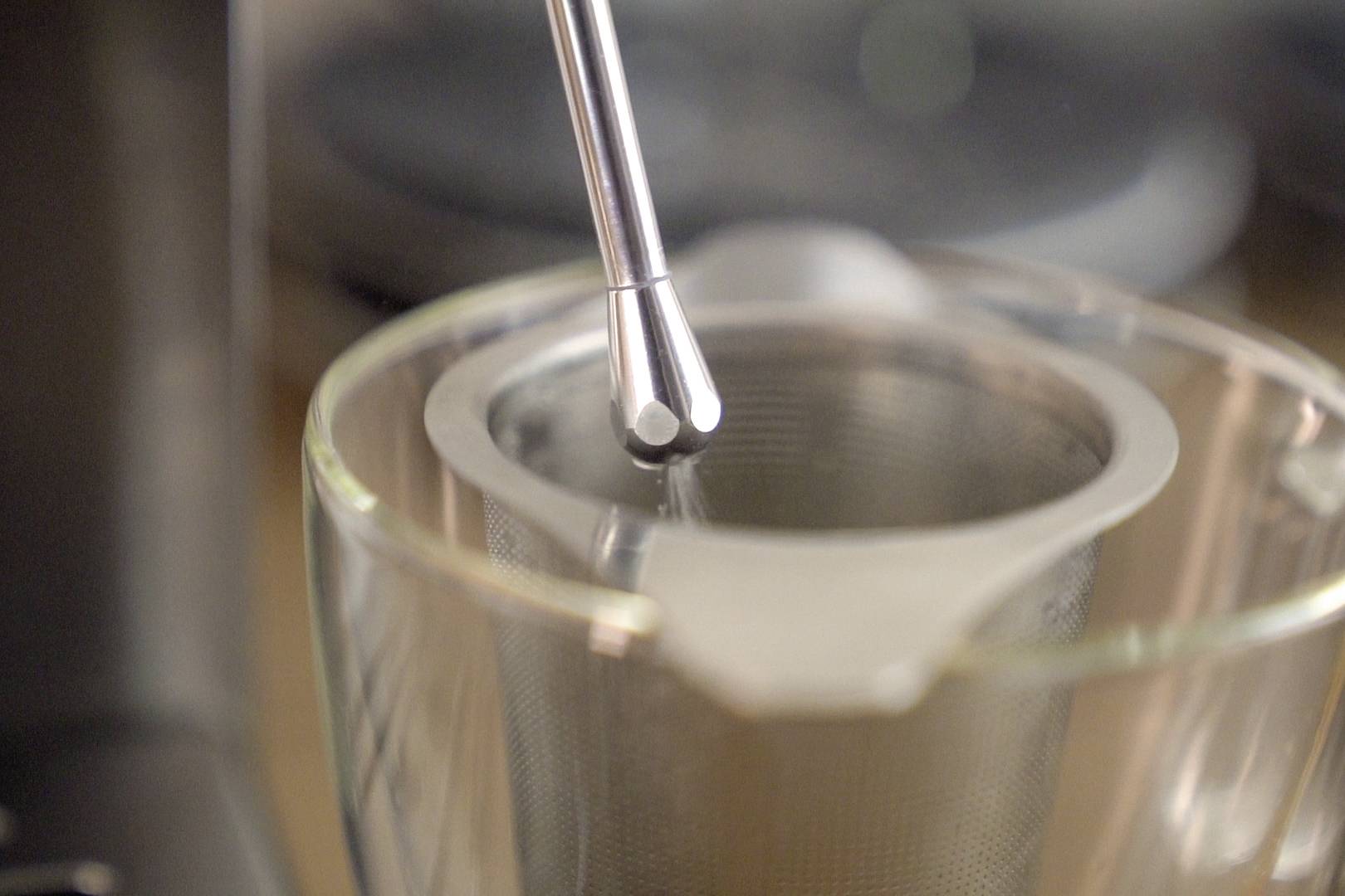 Close-up van een theezeefje in een glazen mok, met een metalen lepel erop. De focus ligt op de bereiding van warme thee.