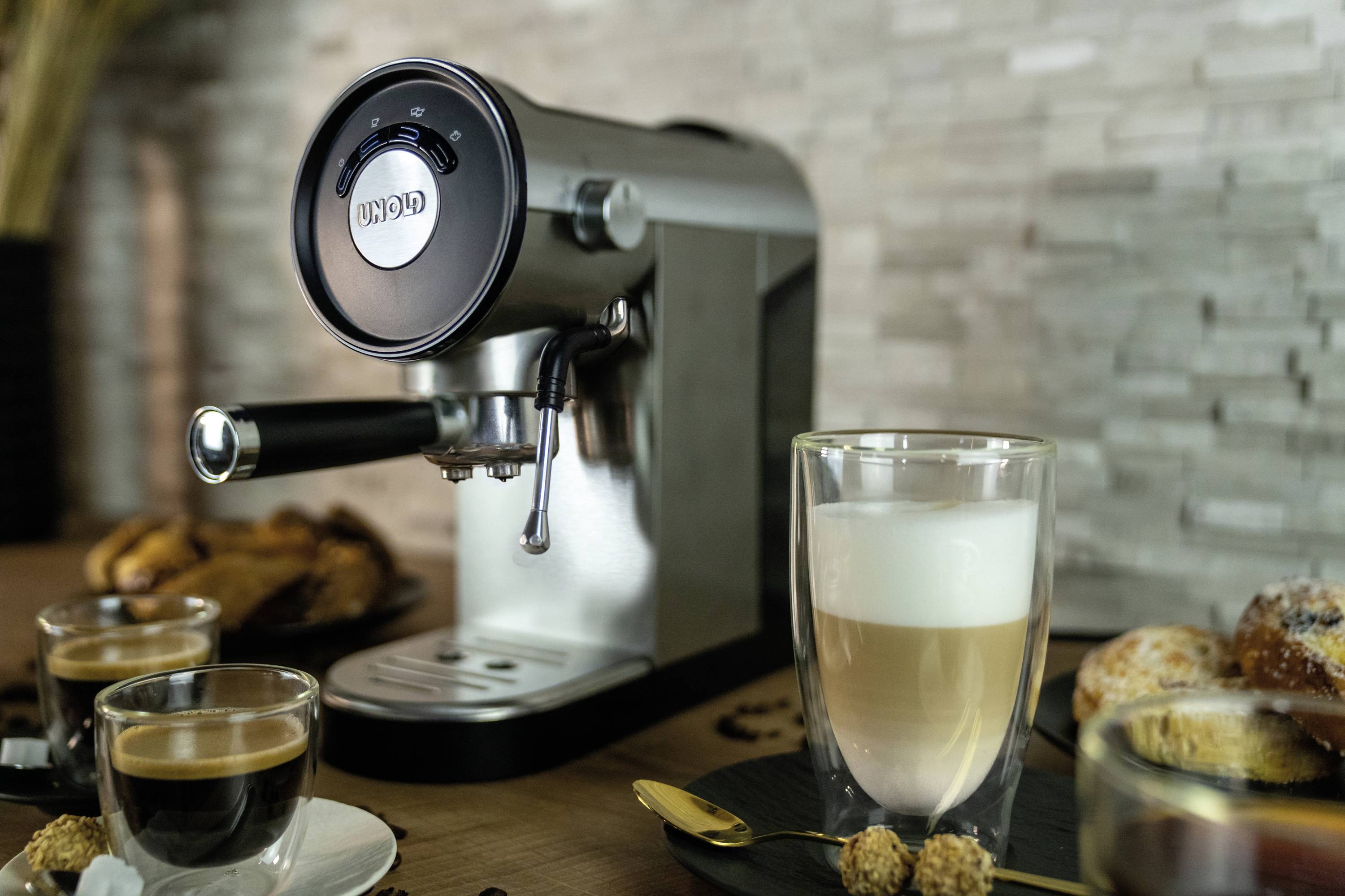 Een zilveren espressomachine op een houten tafel, omringd door twee kopjes koffie en gebak voor een stenen muur.