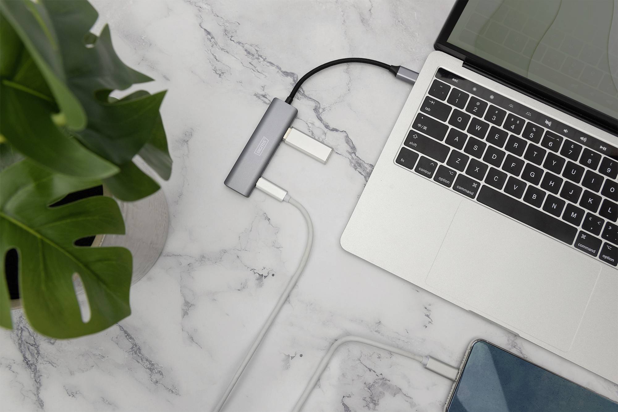Een laptop op een marmeren tafel is via een USB-hub met meerdere kabels verbonden. Ernaast staat een groene plant.