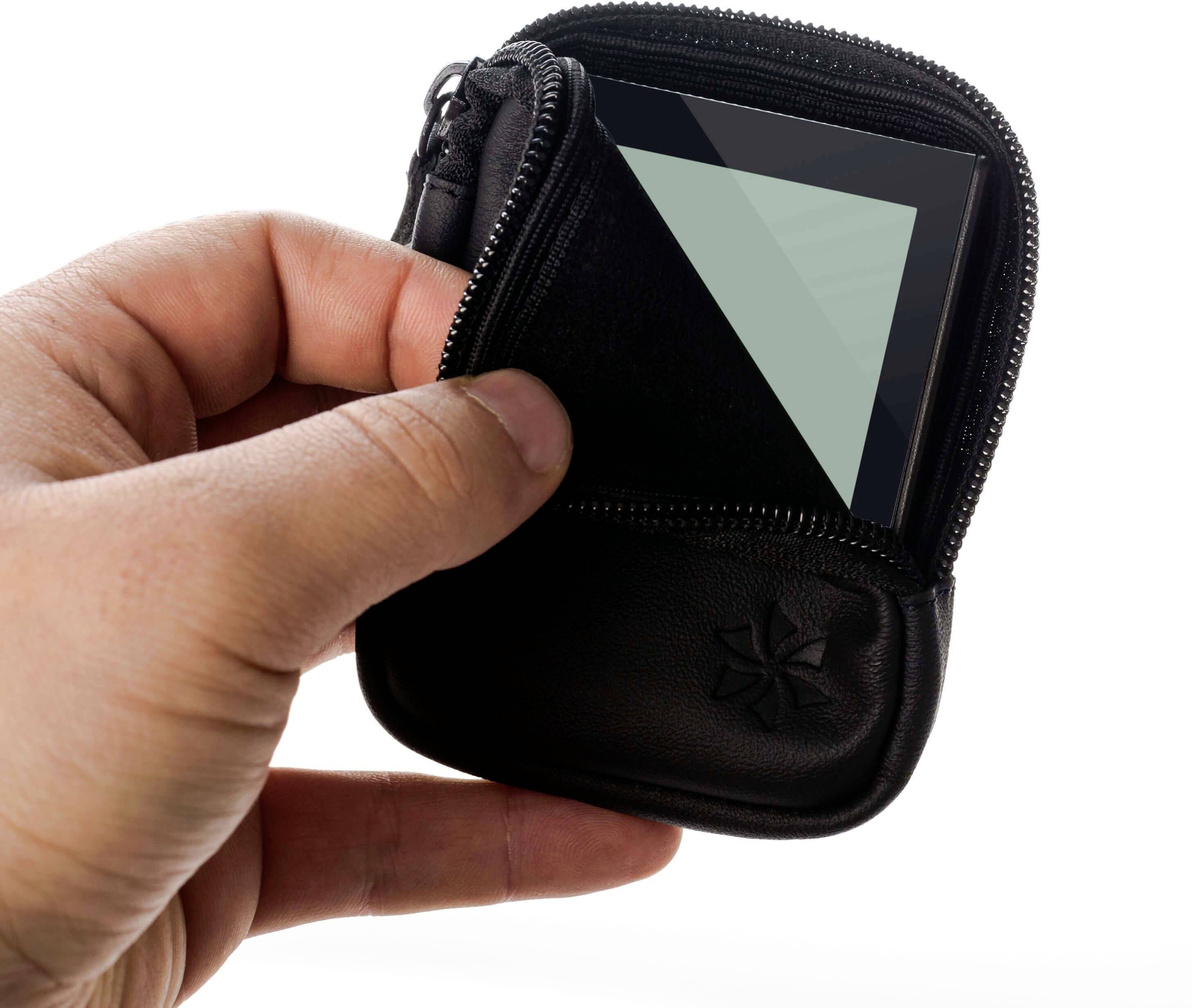 Een hand houdt een kleine zwarte tas met rits vast, waaruit een telefoon gedeeltelijk uitsteekt.
