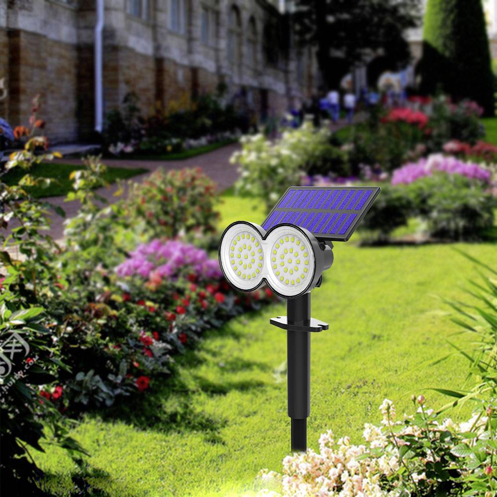Een zonnelamp met twee ronde LED-lampen staat in een zonnige tuin voor bloeiende bloemen en groene struiken.