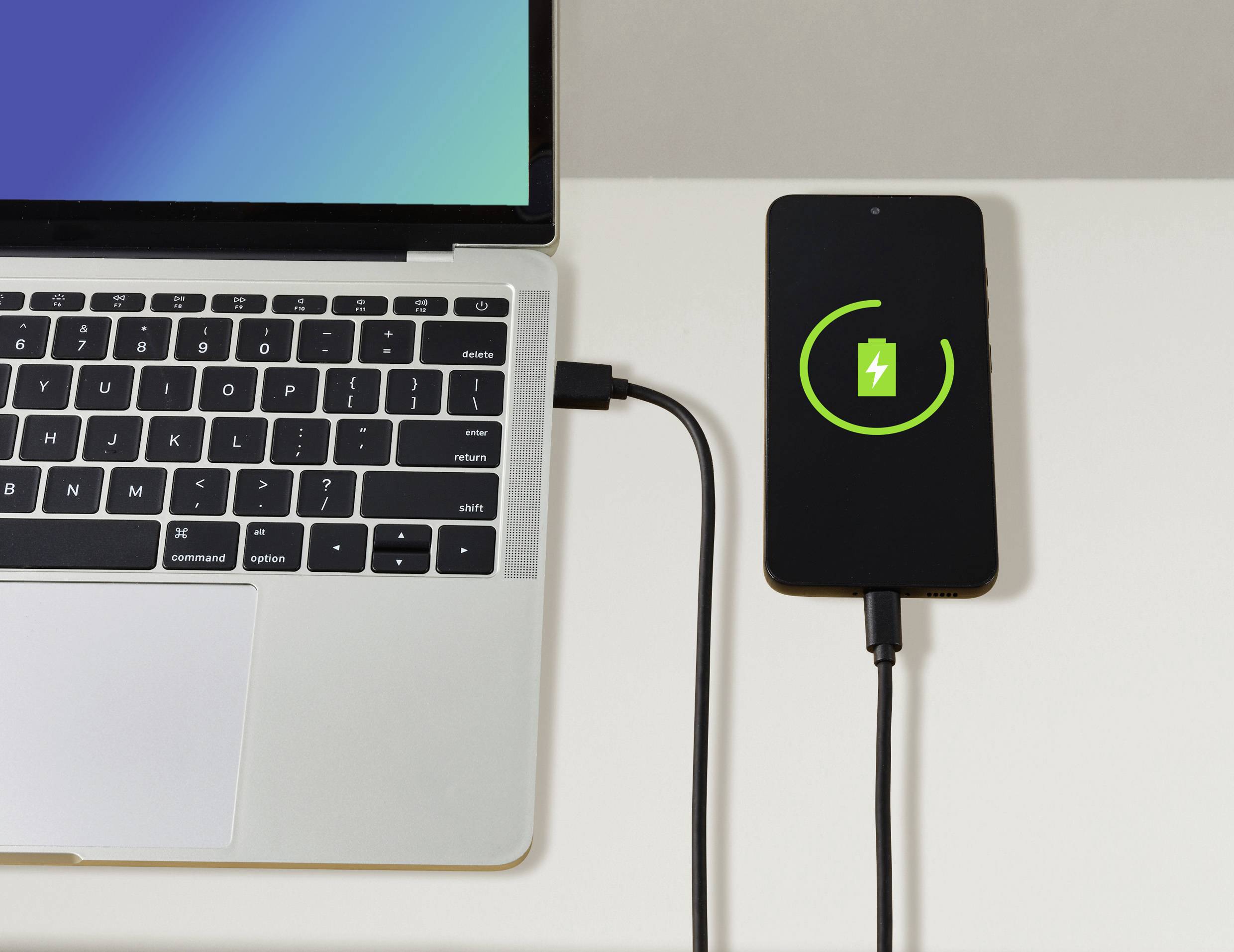 Een smartphone wordt opgeladen via een kabel aangesloten op een laptop. Op het smartphonescherm is een oplaadsymbool zichtbaar.