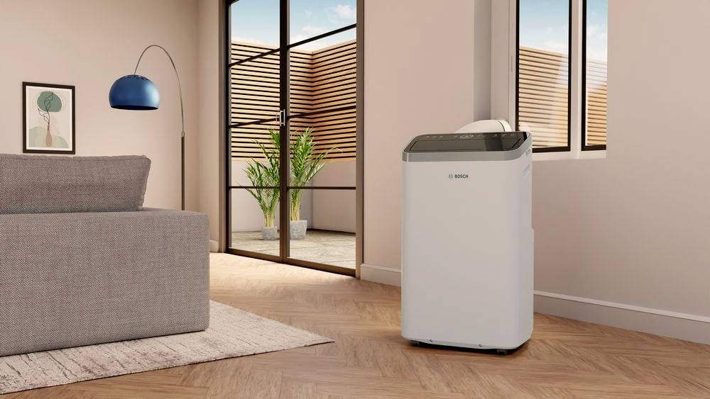 Een draagbare airconditioning staat in een modern woonkamer met houten vloer, naast een grijze bank en grote ramen met uitzicht op een terras.