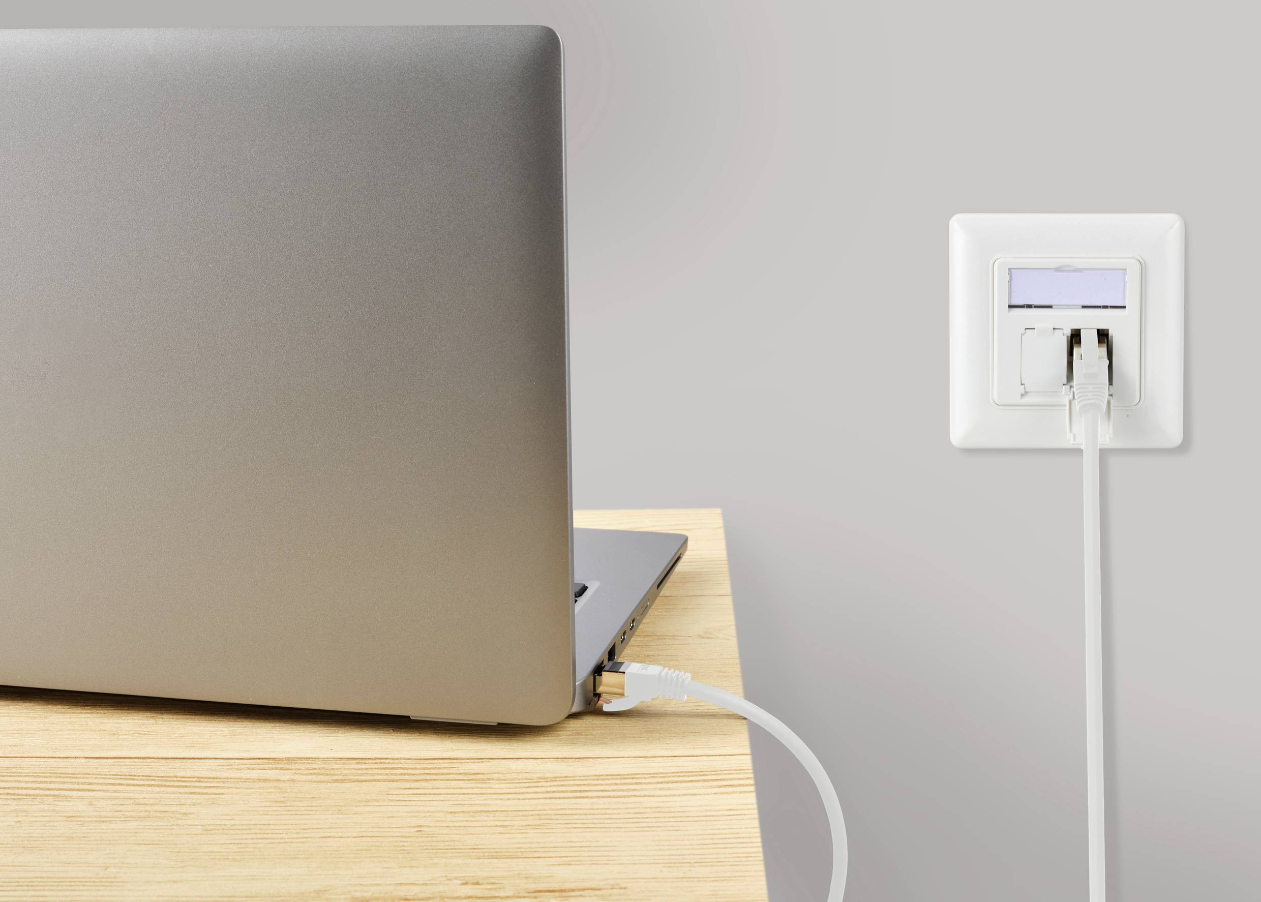 Een laptop op een houten tafel is via een Ethernet-kabel aangesloten op een wandcontactdoos die een internetsignaal doorgeeft.