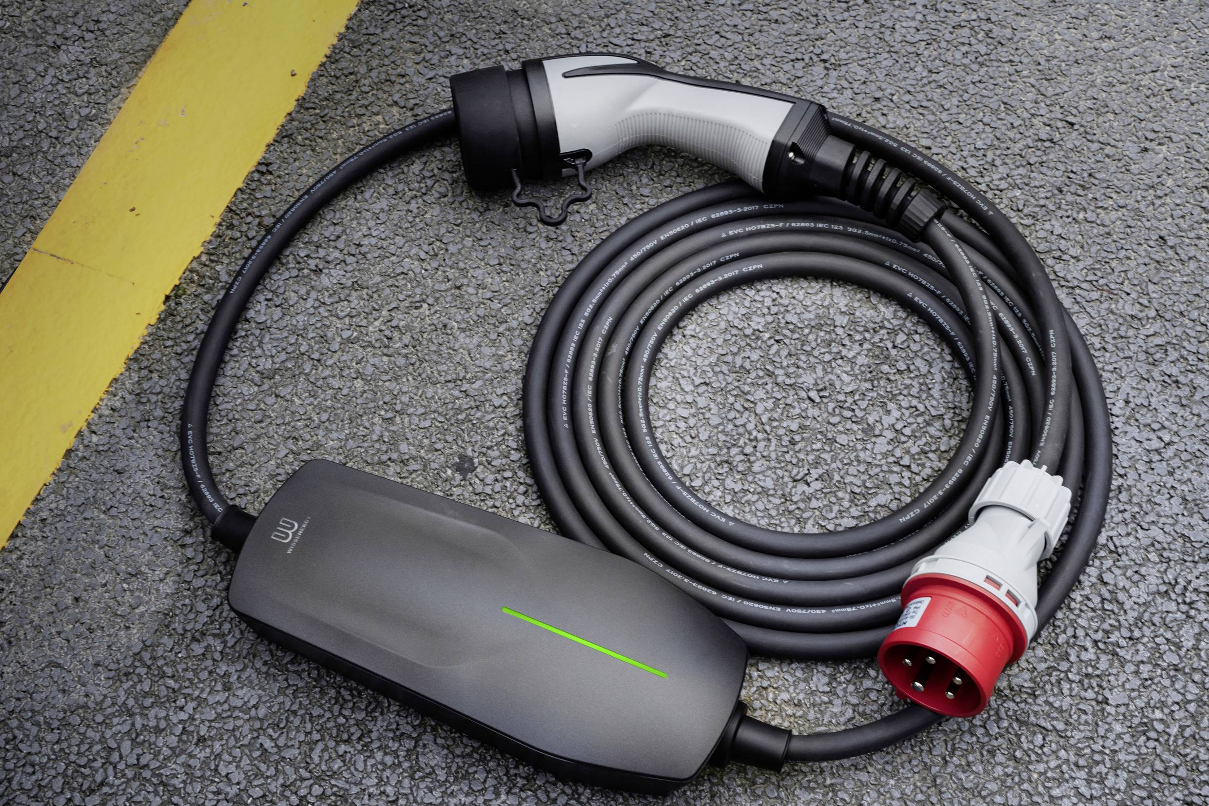 Een grijs oplaadkabel voor elektrische voertuigen ligt op asfalt naast een gele lijn. De kabel heeft een rode stekker.