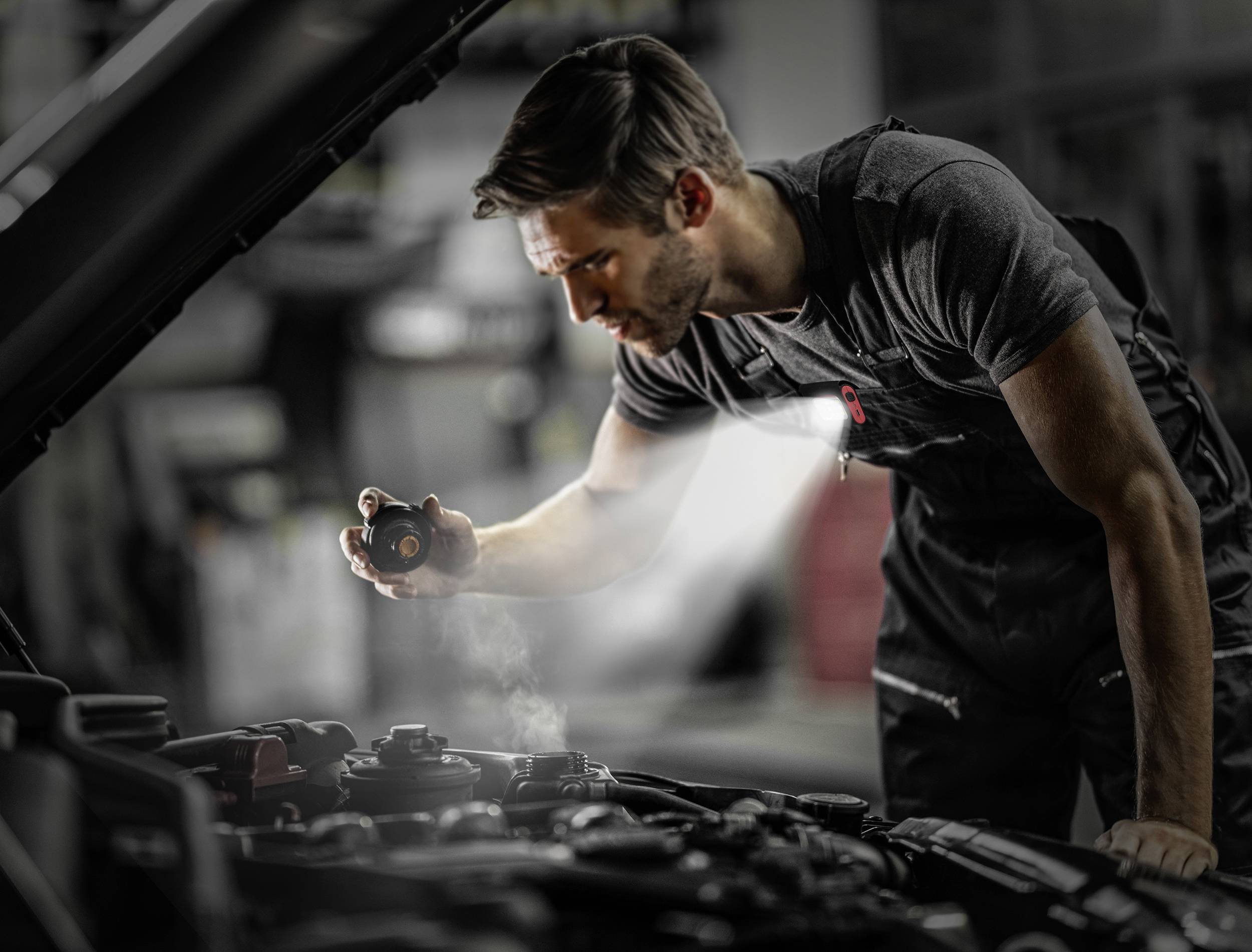 Een monteur inspecteert met een zaklamp de motor van een auto in een werkplaats. De focus ligt op de geconcentreerde werkzaamheden.