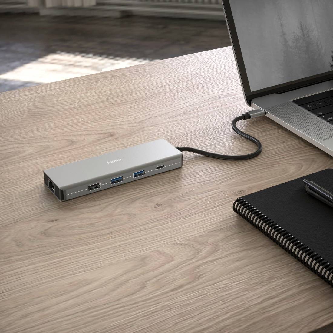 Een laptop staat op een houten tafel naast een notitieboek. Aangesloten is een USB-hub met meerdere aansluitingen, waaronder USB en HDMI.