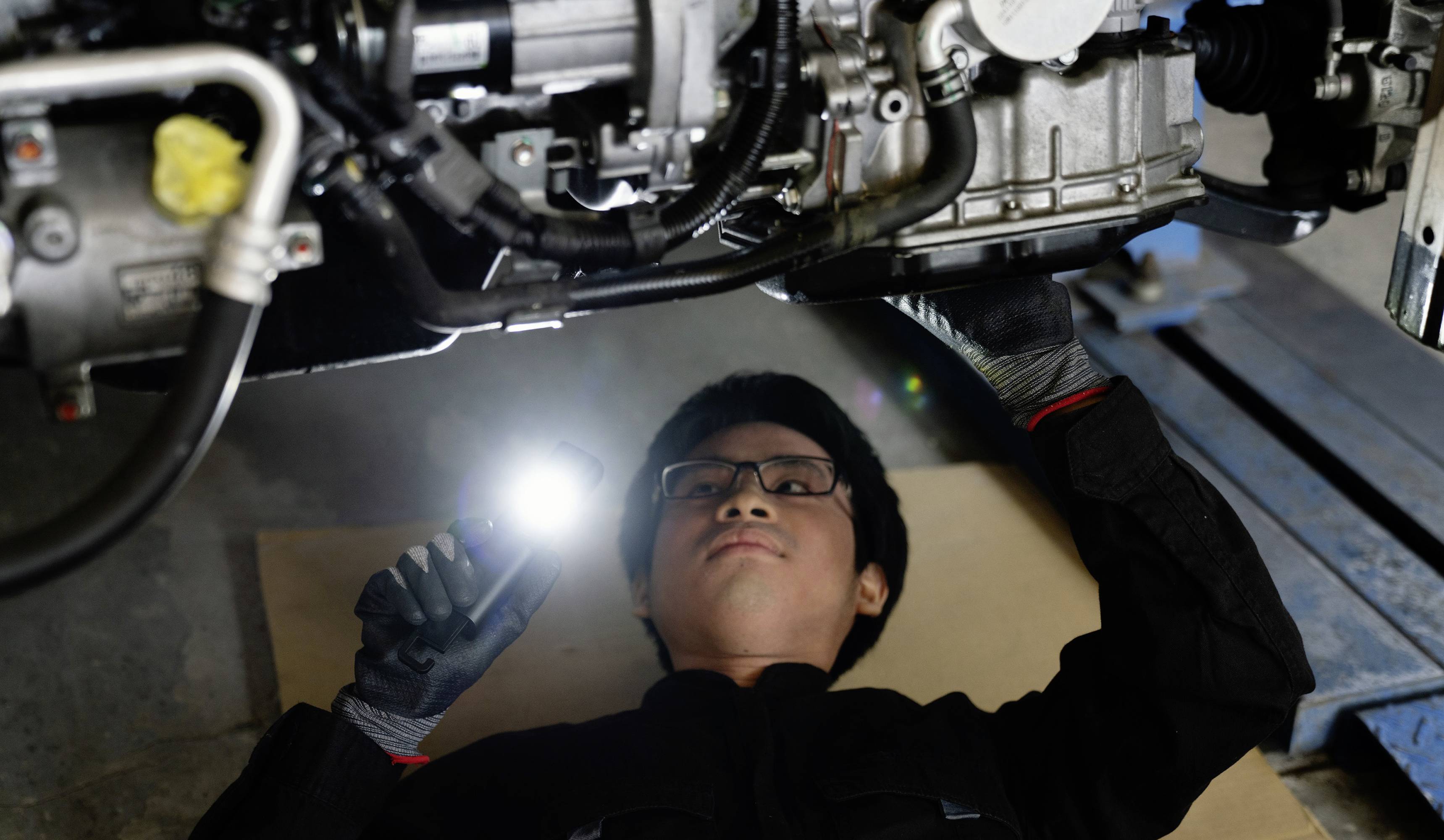 Een persoon ligt onder een auto en inspecteert de motor met een zaklamp. Ze draagt handschoenen en een veiligheidsbril.