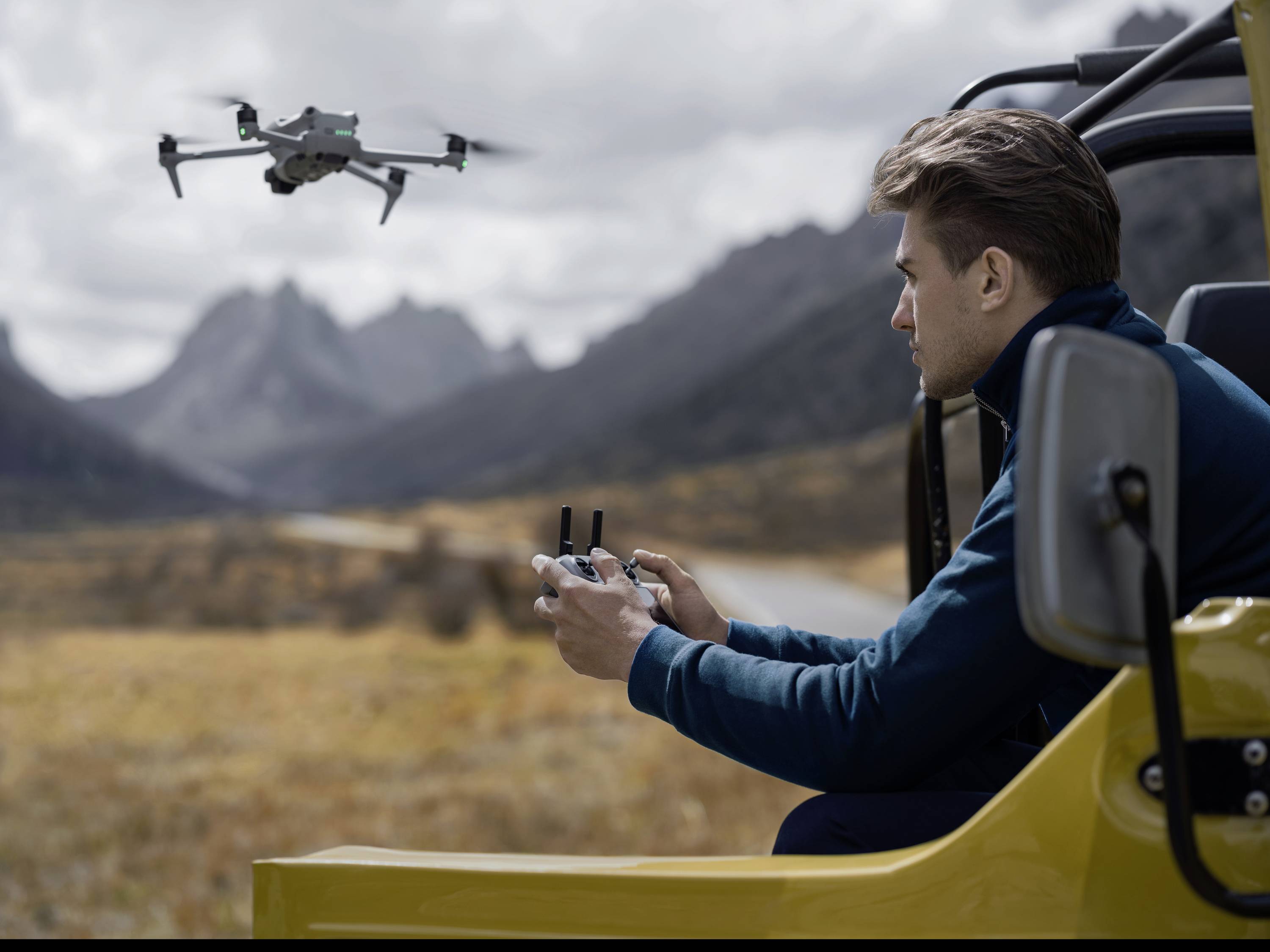 Een man bestuurt een drone vanuit een open terreinwagen. Op de achtergrond een berglandschap. De drone vliegt over grasland.