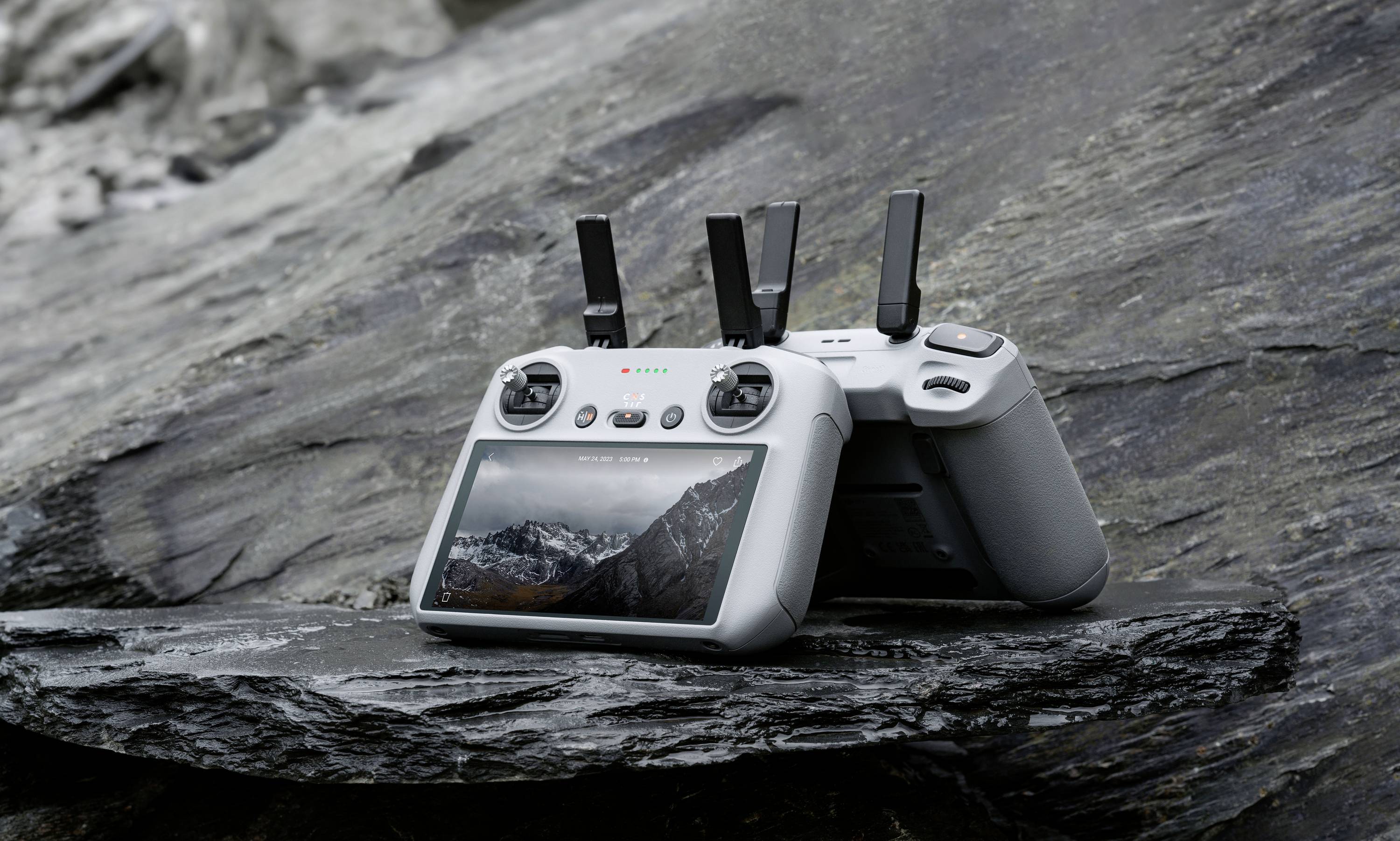 Een grijze drone-afstandsbediening met een display waarop een berglandschap te zien is, ligt op een rots in de buitenlucht.
