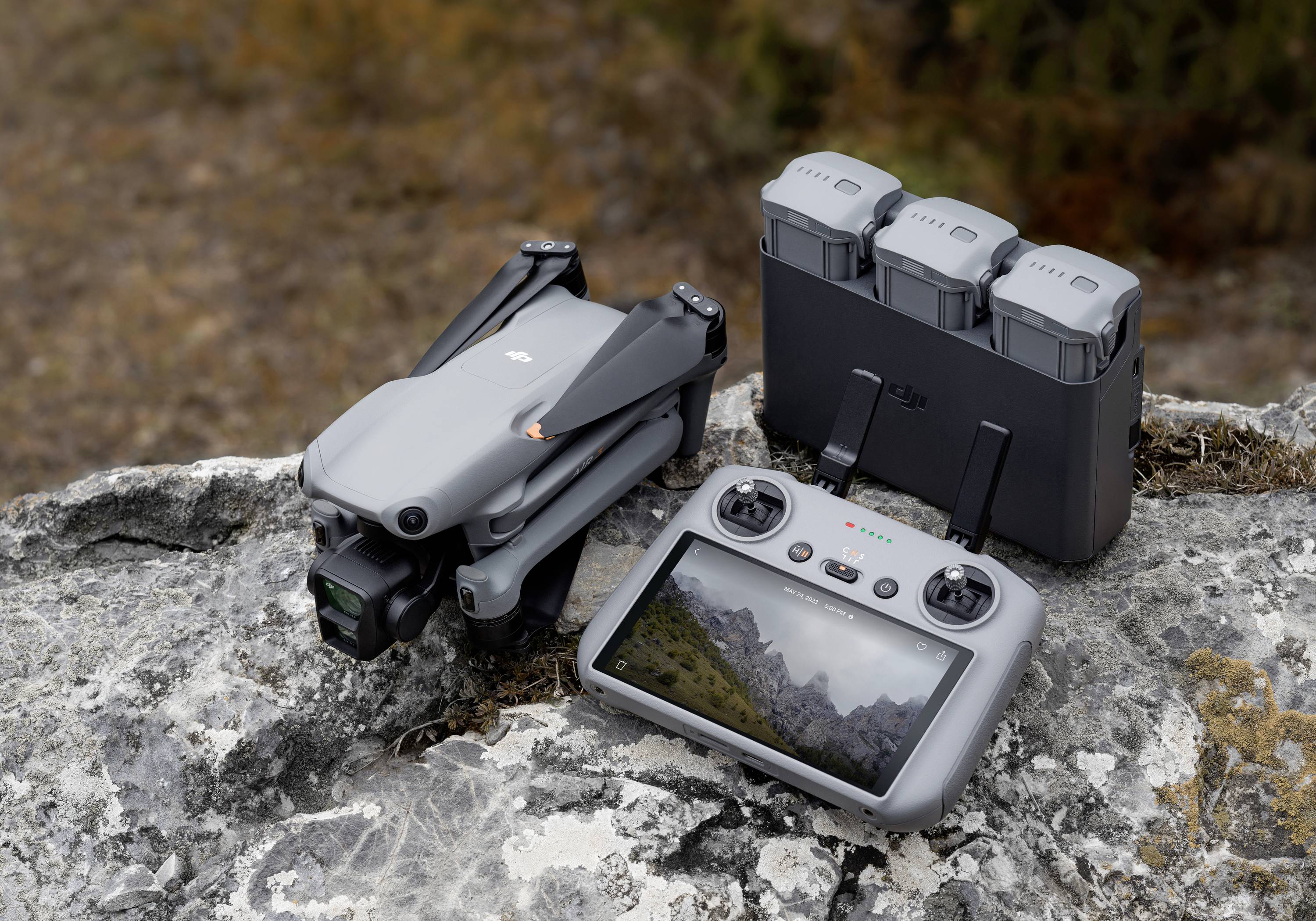 Een grijze drone en een afstandsbediening met een berglandschap op het display liggen op een rots. Ernaast een oplader met drie accu's.