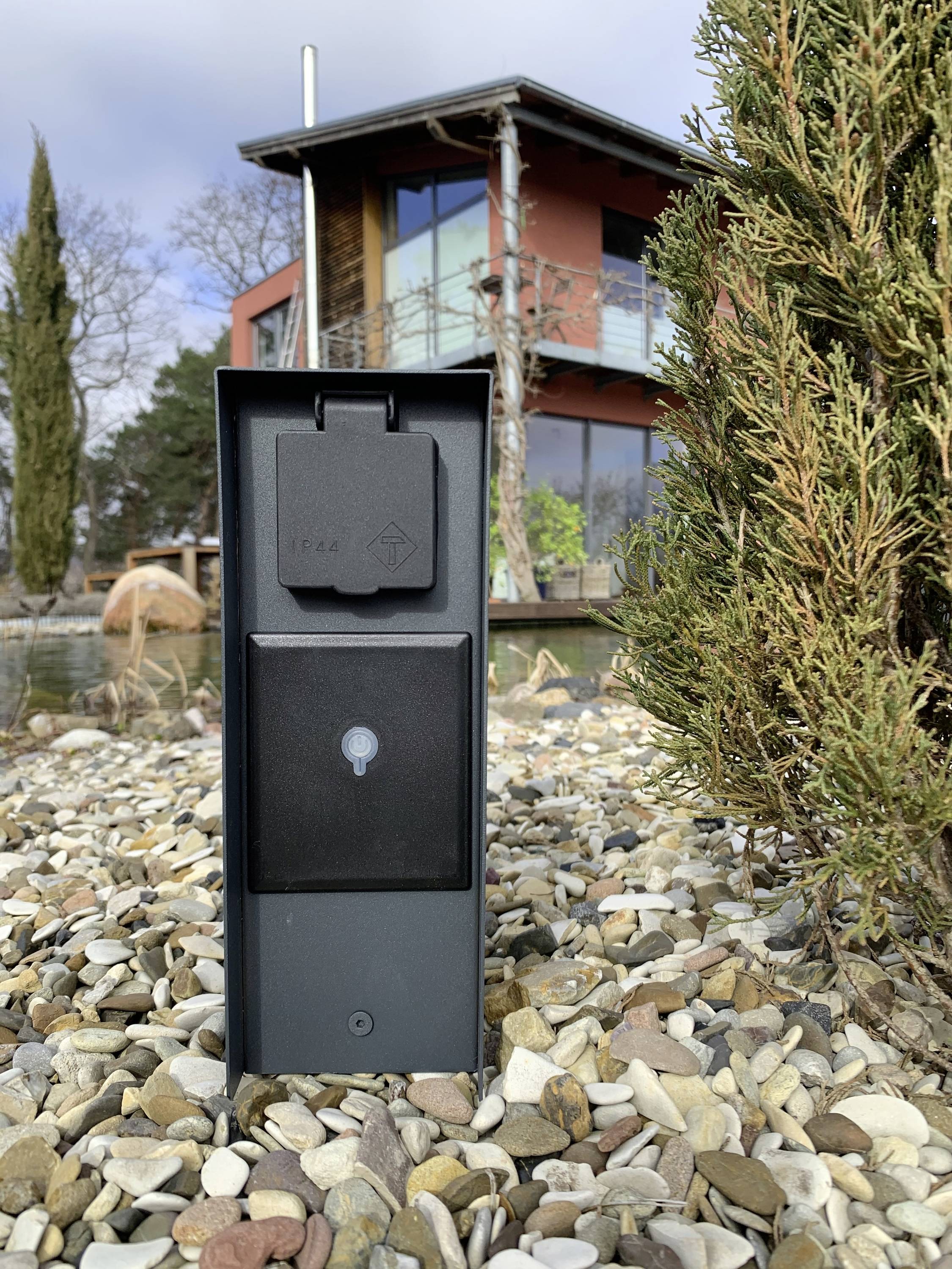 Een moderne buitenstopcontactdoos staat op een stenige ondergrond, met een huis op de achtergrond en een boom rechts in het beeld.